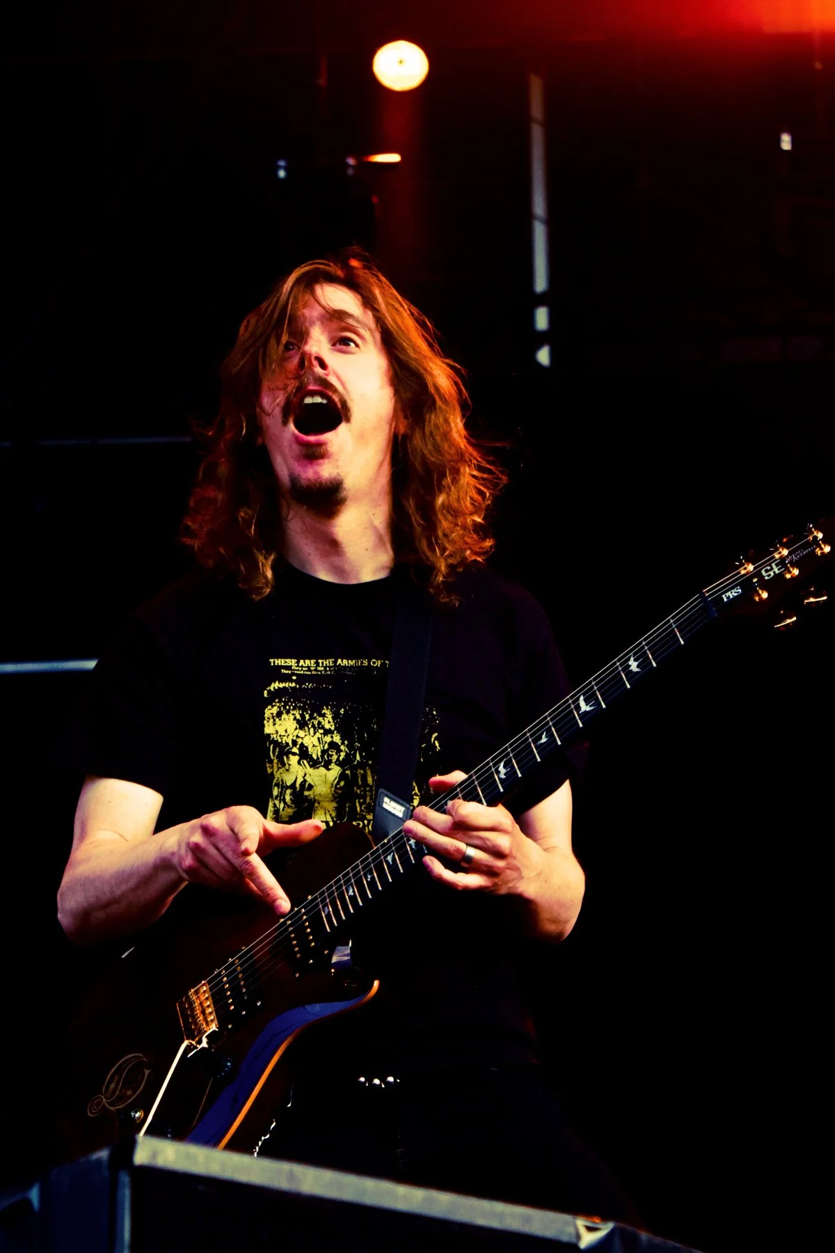 A man with long curly hair and a mustache, singing passionately while playing an electric guitar on stage, illuminated by stage lights.