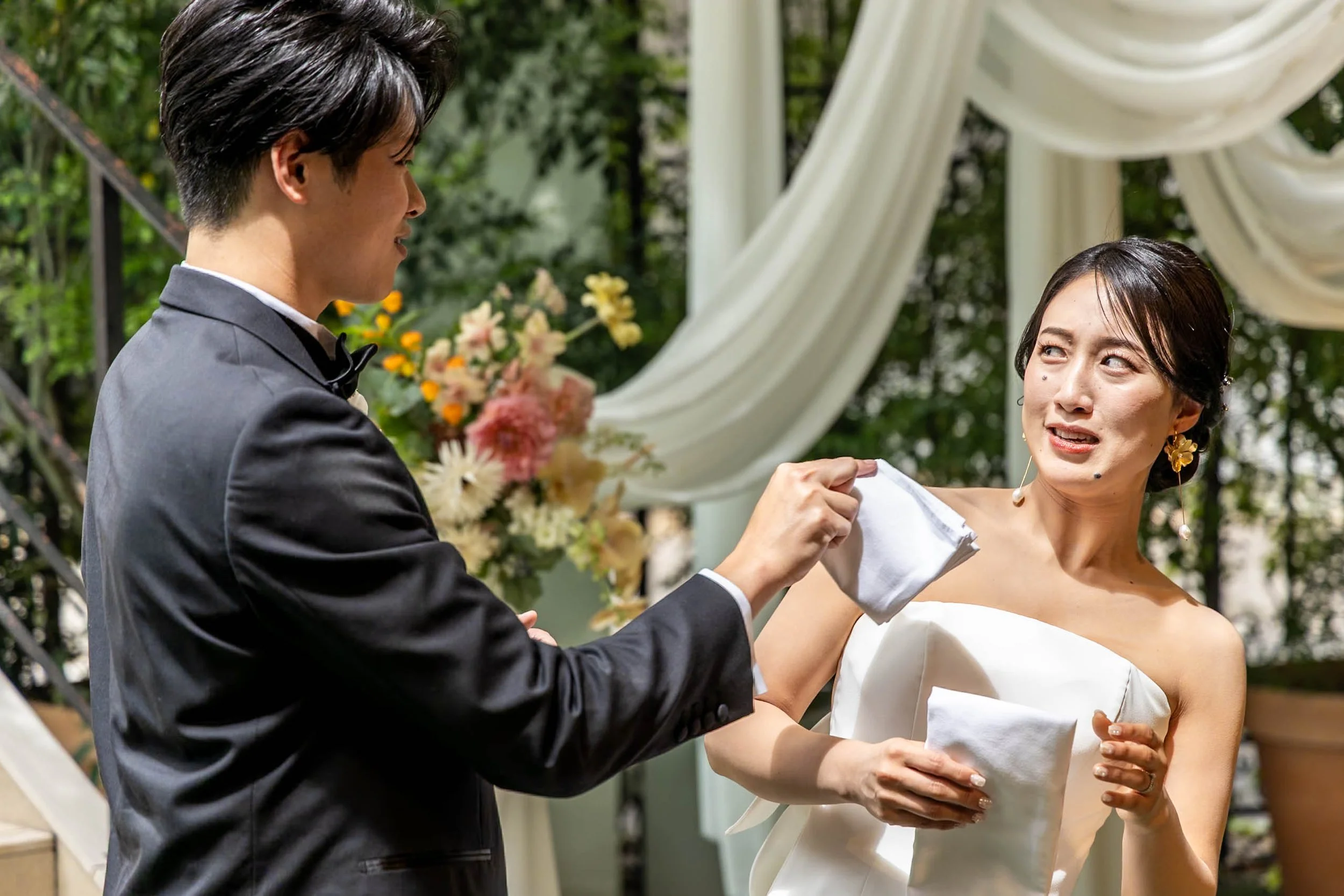 A man in a tuxedo is holding a tissue to a woman in a wedding dress, who appears emotional, during a wedding ceremony outdoors with floral decorations and white drapery.