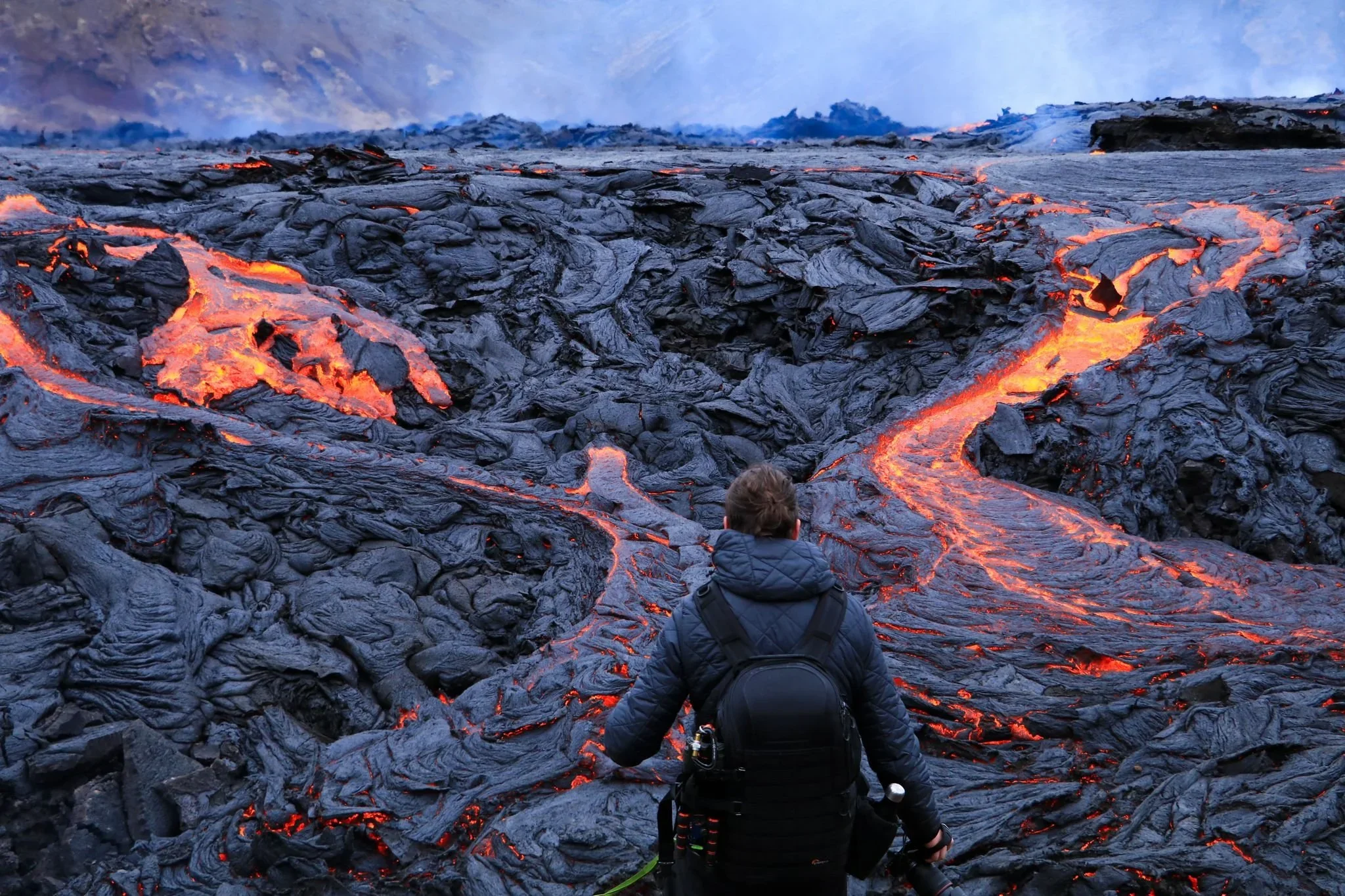 A person with a backpack and camera stands amidst glowing lava flows on a volcanic landscape.