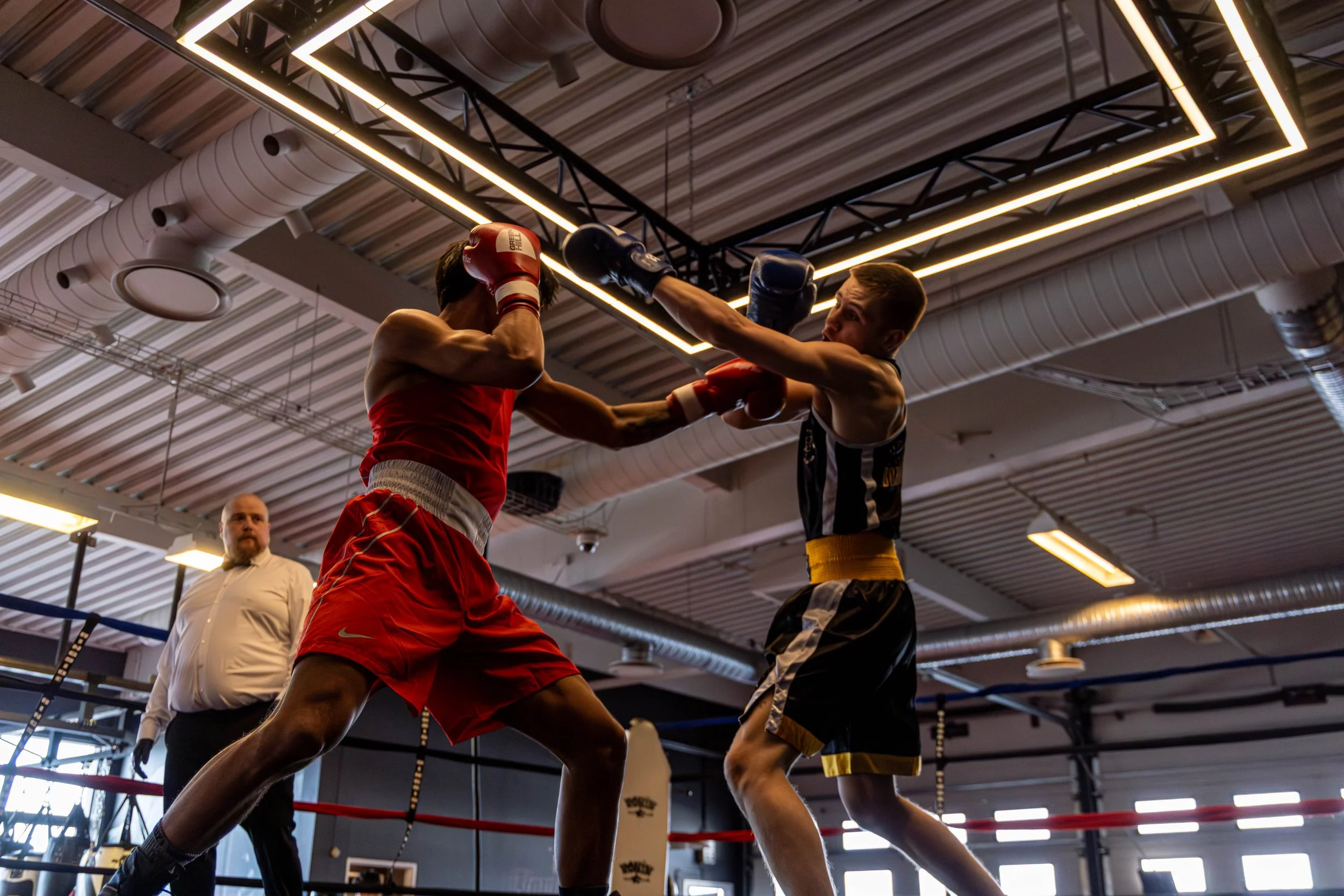 Two male boxers sparring in a ring, one in red shorts and the other in black and white shorts, with a referee observing in the background, inside a boxing gym with modern lighting fixtures.
