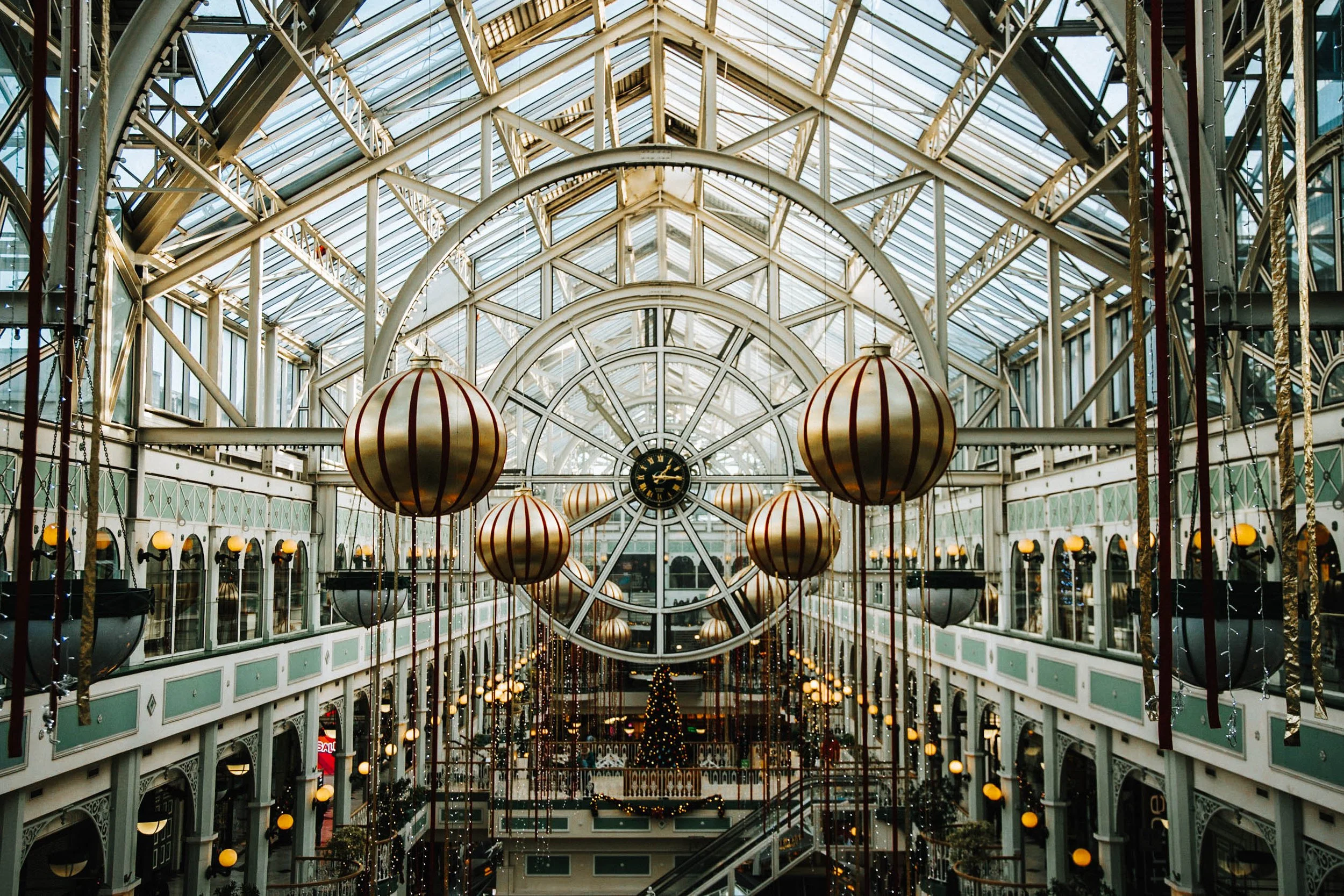 Interior view of a grand shopping mall with festive Christmas decorations, including large gold and red striped ornaments hanging from the ceiling and a decorated Christmas tree in the background. The mall features a glass ceiling and upper balconies