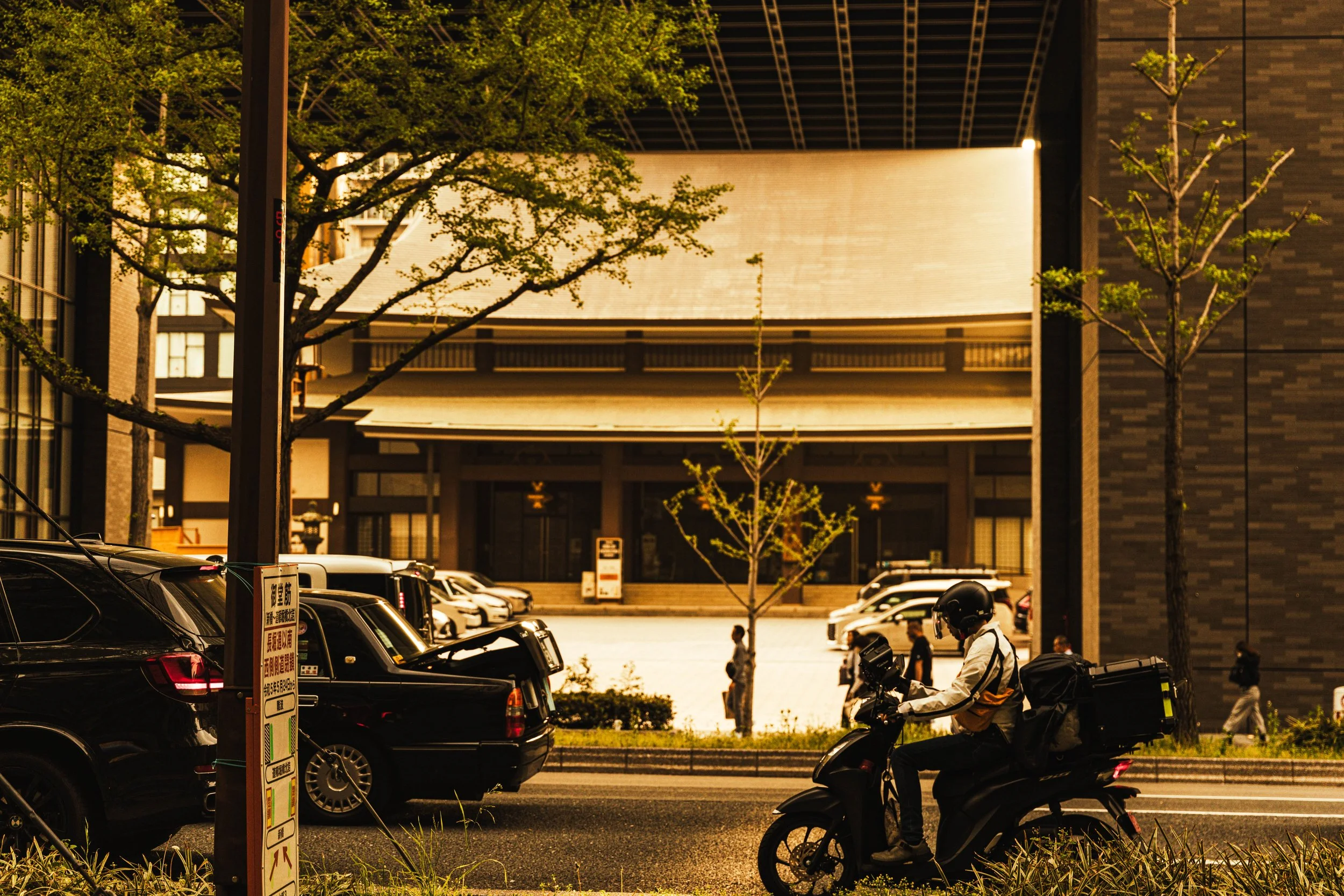 A person riding a scooter on the street, wearing a helmet and a white jacket, with a large dark building and parked cars in the background, and trees lining the street.