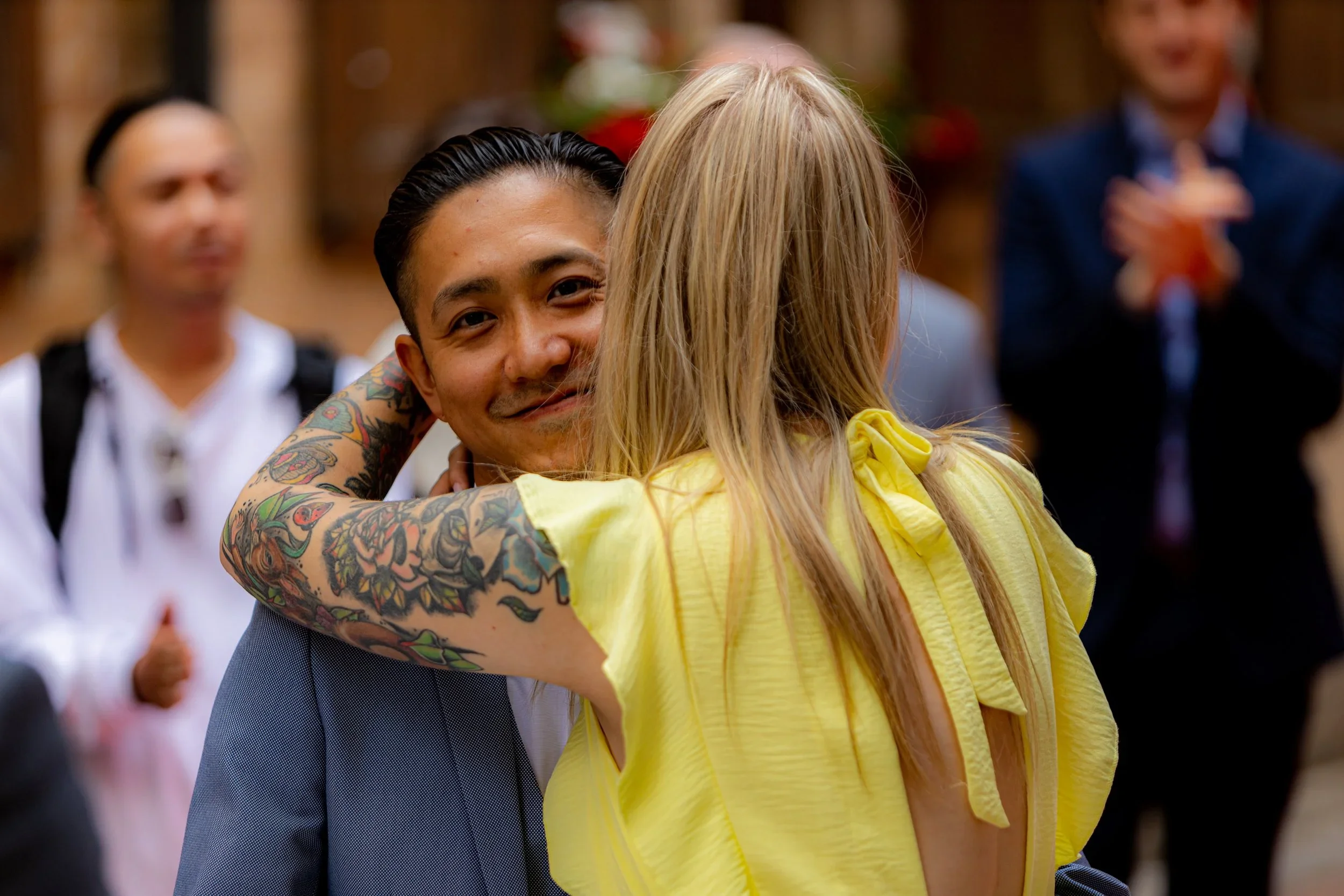 A man with tattoos smiling as he is embraced by a woman with blonde hair wearing a yellow dress, at a social gathering with other blurred people in the background.