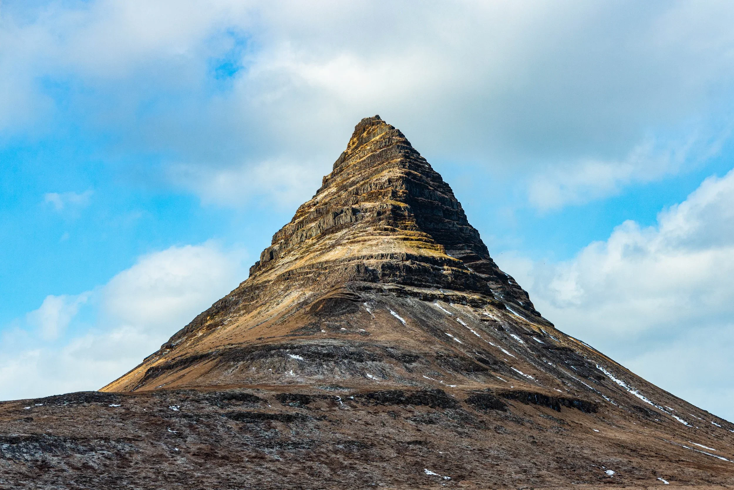 A mountain with a conical, layered shape against a partly cloudy sky.