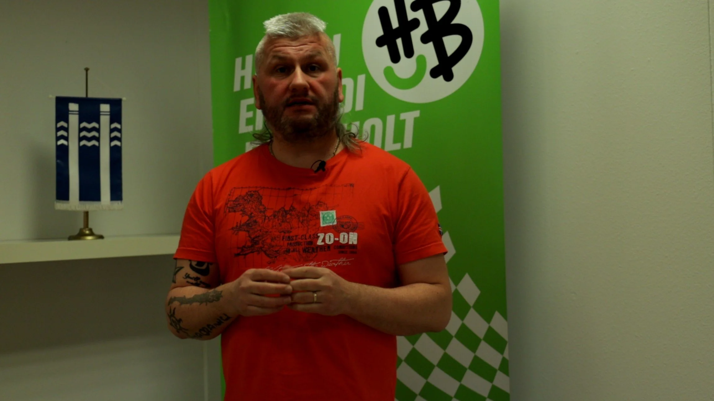 A man with a mullet hairstyle and tattoos on his arms, standing in front of a green backdrop with cryptocurrency symbols, wearing a red T-shirt with a graphic design.