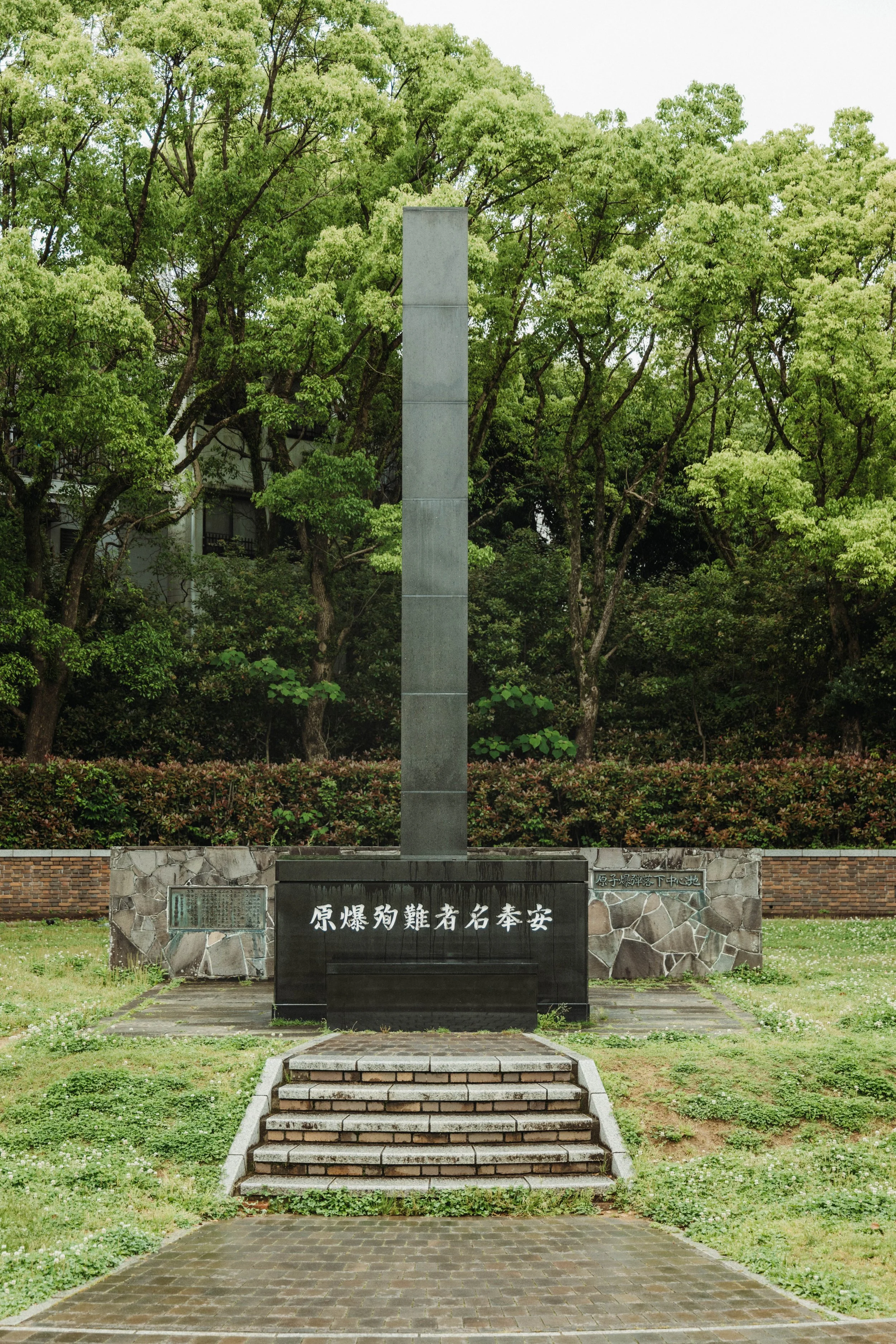 A memorial monument with a tall rectangular pole on a stone and brick base, surrounded by greenery and trees.
