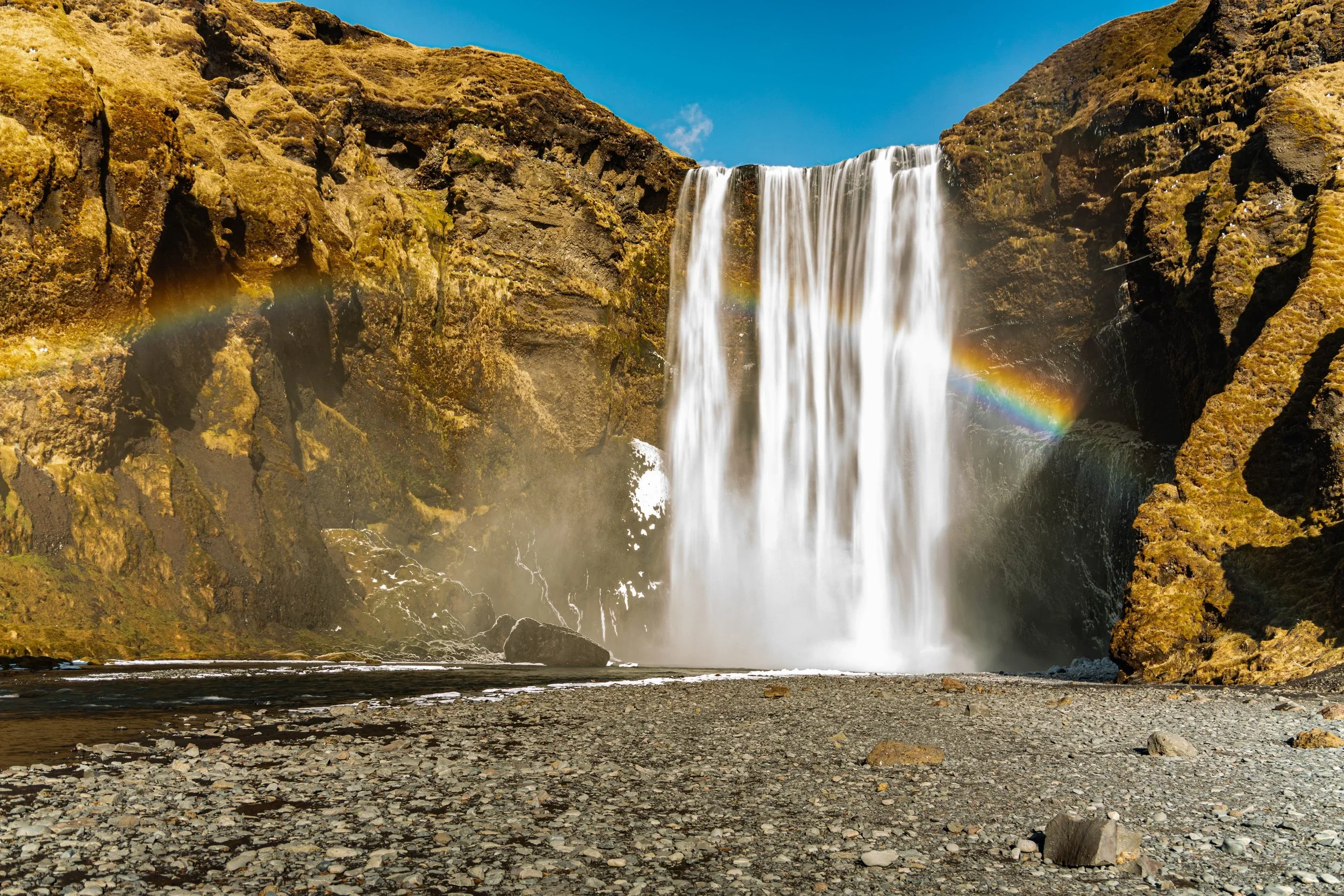 A tall waterfall cascading down a rocky cliff, with a rainbow visible in the mist to the left and right of the waterfall, under a clear blue sky.