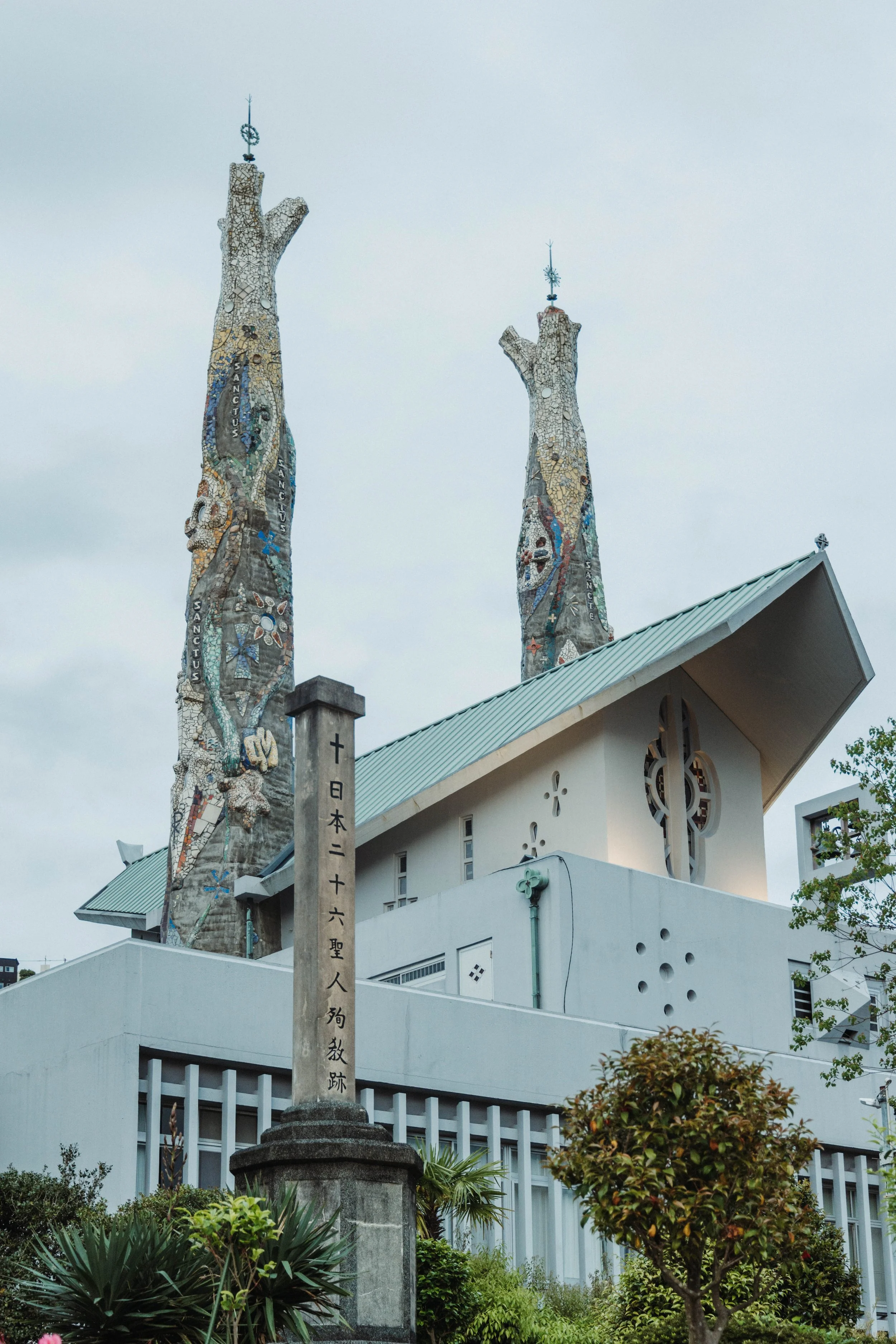 The image shows a modern church building with a sloped roof, featuring unique circular window designs. Behind it, two tall, twisted towers decorated with colorful mosaic patterns and religious symbols are visible, topped with metal spires. In the for