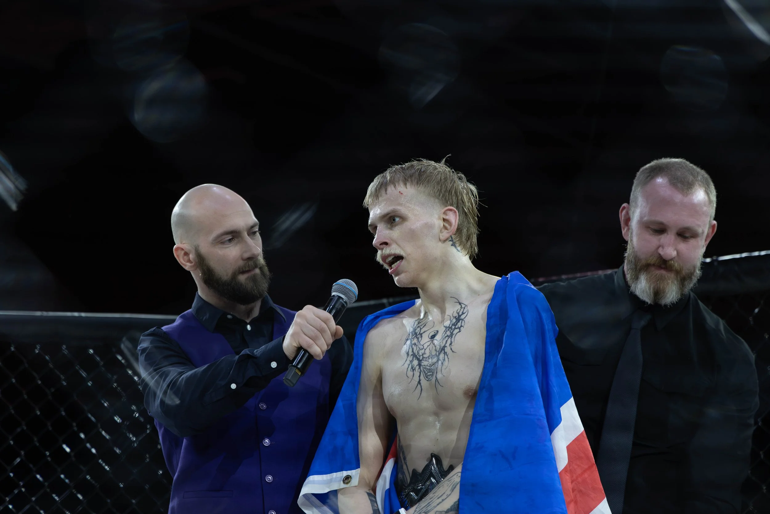Mixed martial arts fighter with tattoos and a flag draped over their shoulders, speaking into a microphone, flanked by two men in black shirts, inside an MMA cage.