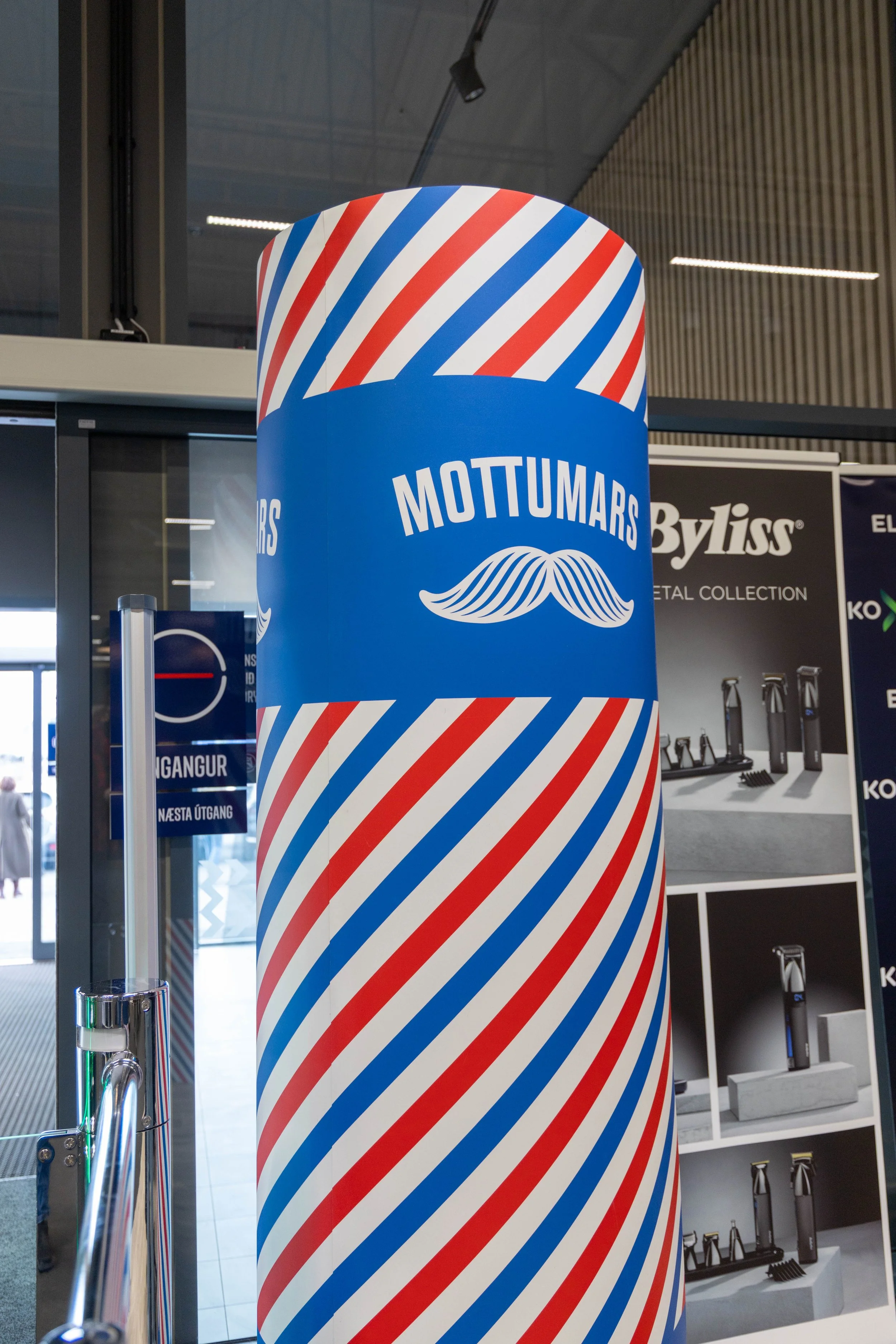 A large cylindrical display or sign with red, white, and blue diagonal stripes, featuring the logo of Mottumars, which includes a stylized mustache and the brand name in white text.