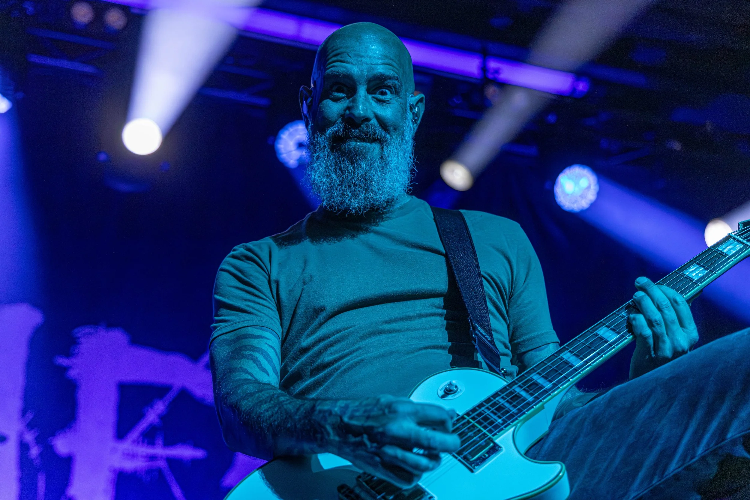 A bearded man with a bald head playing an electric guitar on stage during a concert with colorful stage lighting.
