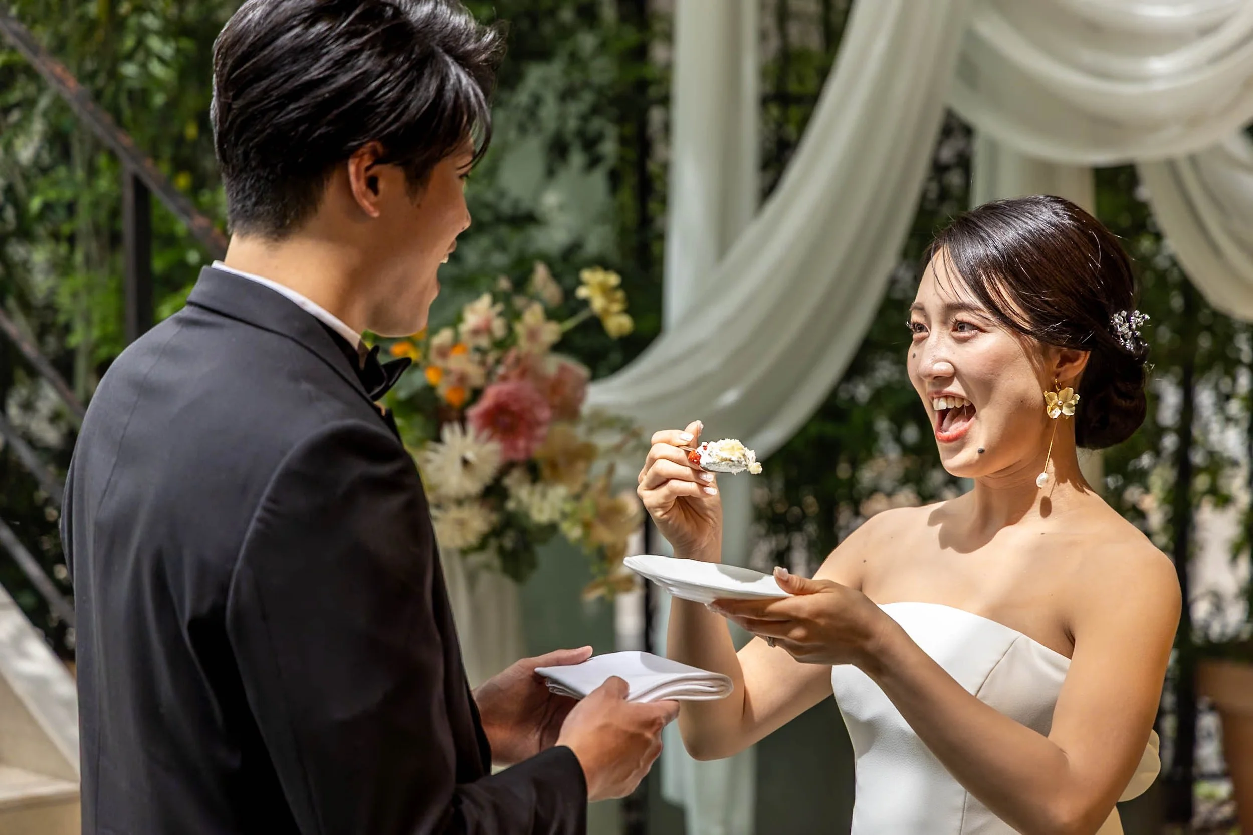 A bride in a white wedding dress and a groom in a black suit are exchanging wedding vows outdoors. The bride is smiling and holding a piece of cake, while the groom is holding a napkin and smiling. There are flowers and a draped white fabric in the b