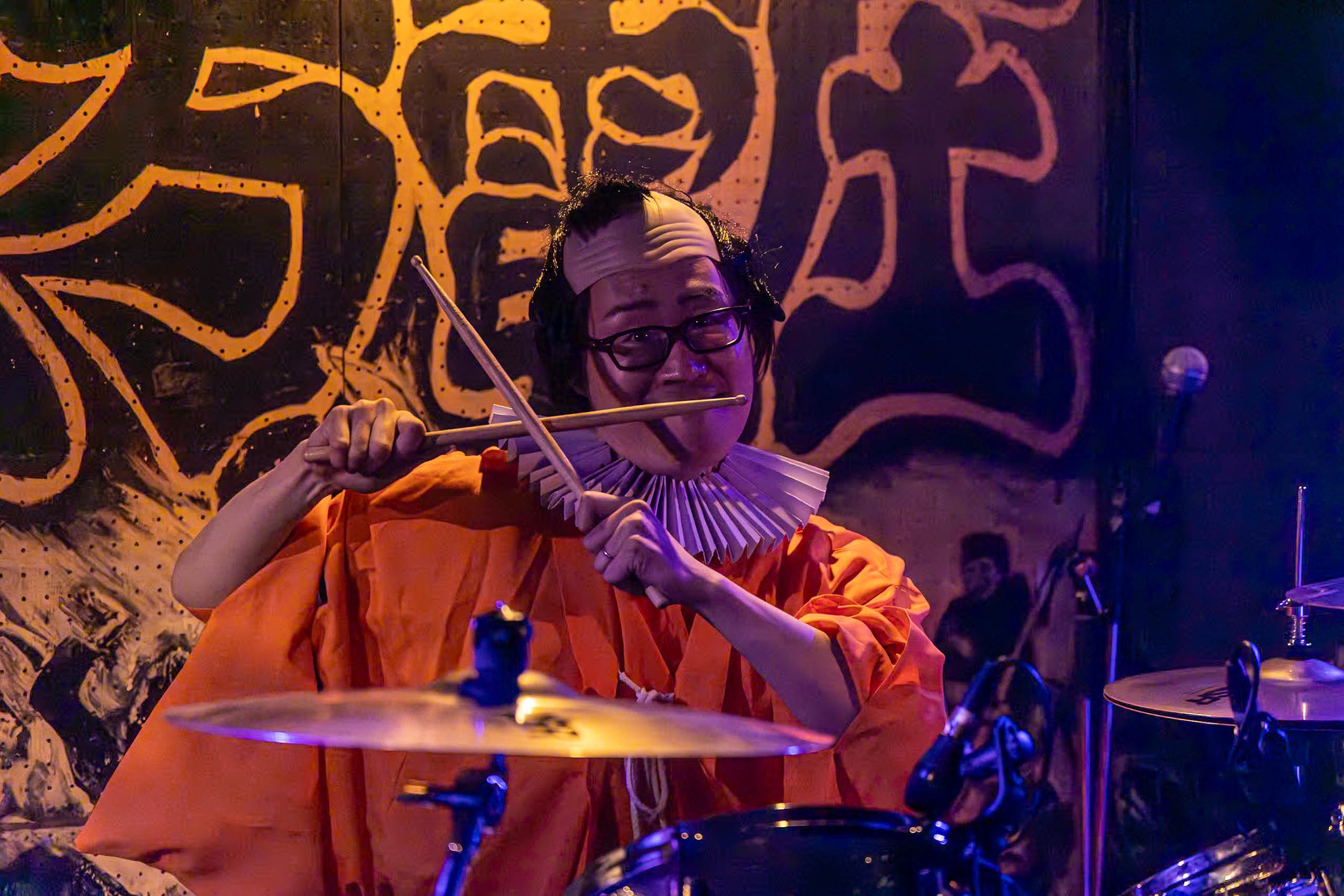 A musician dressed in orange with a fan collar, playing drums and holding drumsticks in a colorful, artistic setting with wall art in the background.