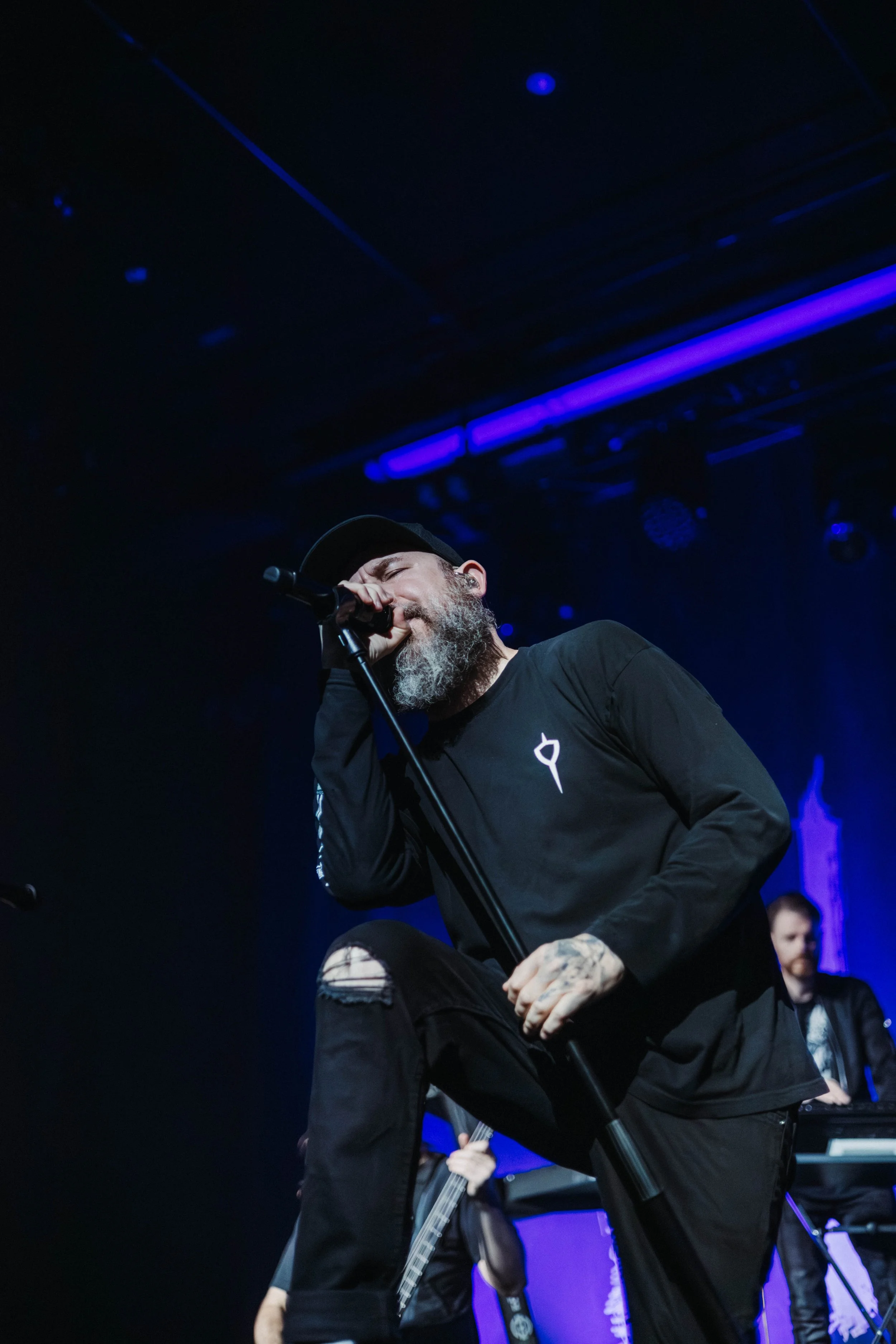 A male singer with a beard and tattoos, wearing a black hat and black clothing, performs on stage with a microphone in hand, surrounded by band members with musical instruments in a concert setting with blue stage lights.