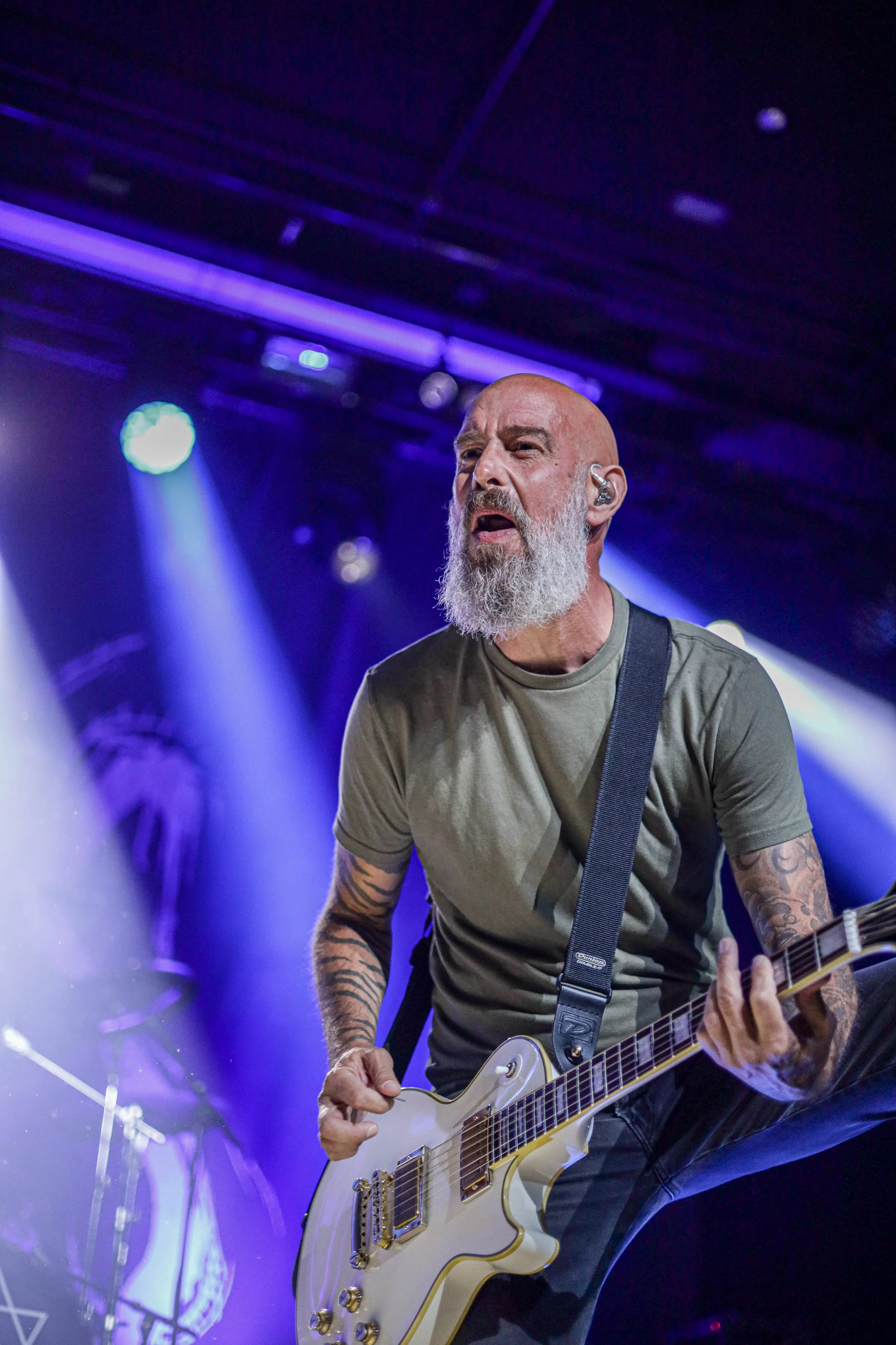 A bearded man with tattoos wearing a gray t-shirt and playing an electric guitar on stage under purple and blue lights.