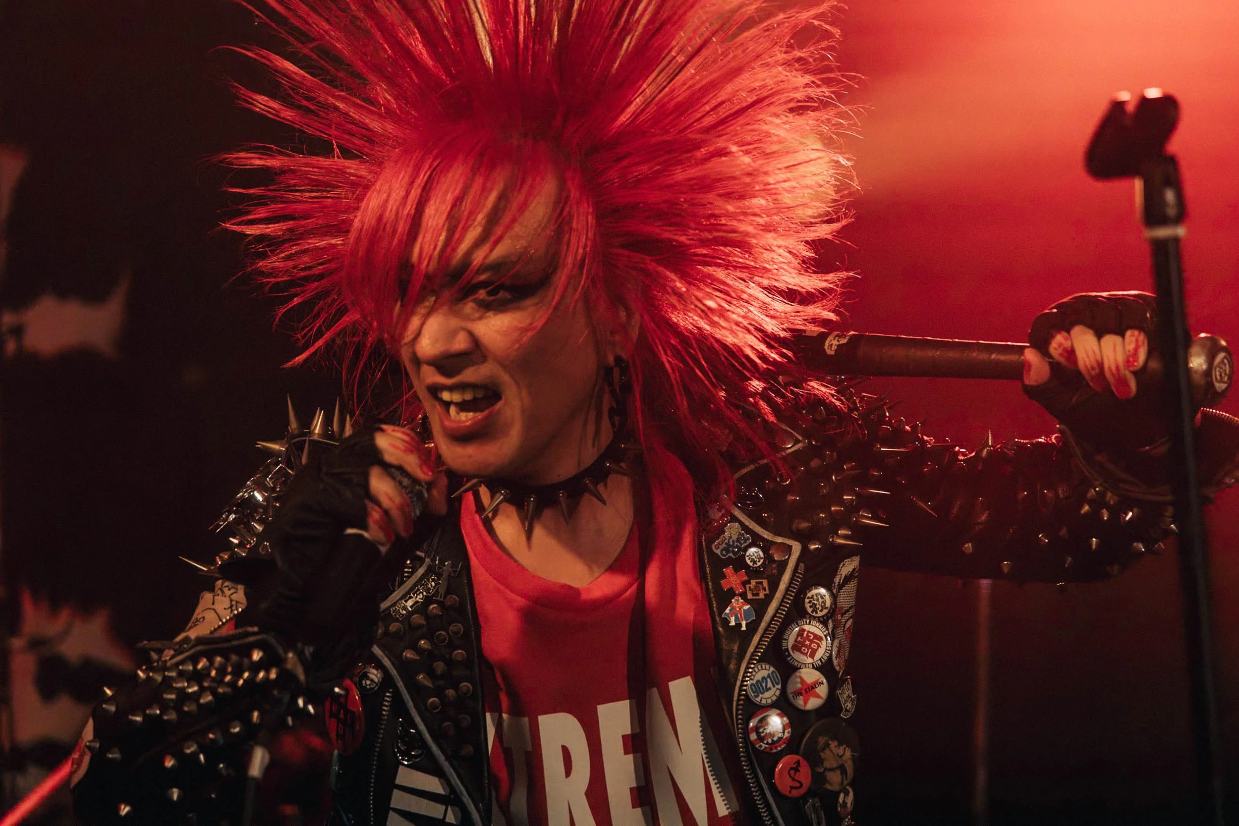 A punk musician with bright red spiked hair and a spiked leather jacket, performing passionately on stage.
