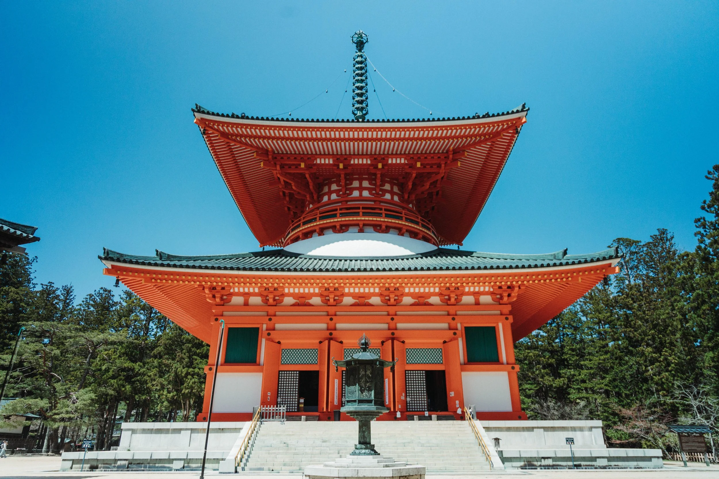 Traditional Japanese temple with a multi-tiered pagoda, red and white walls, dark roof tiles, and surrounded by green trees under a clear blue sky.