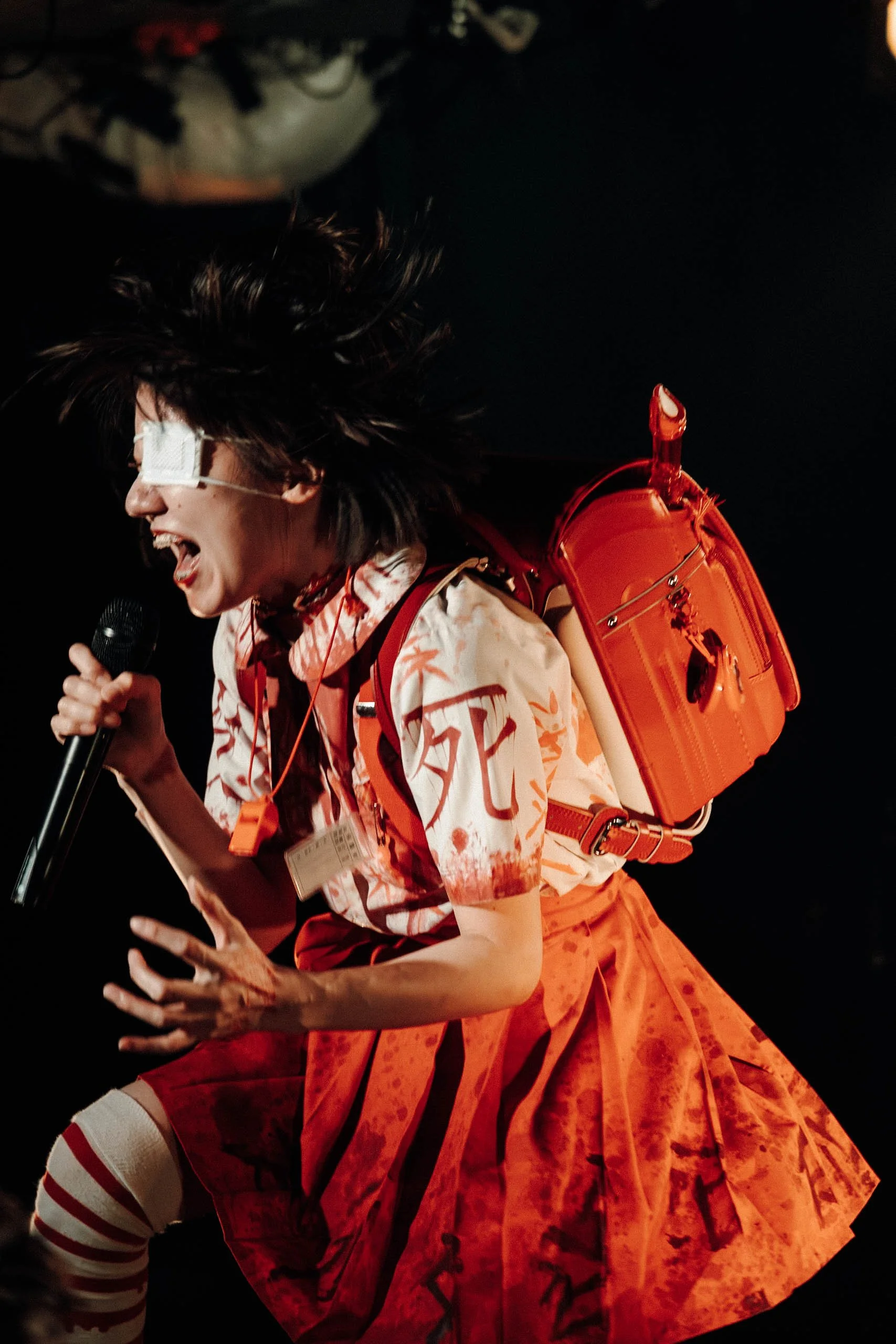 A person with unkempt hair, wearing an eye patch, a patterned shirt, and a red skirt, holding a microphone and leaning forward on stage with a bright orange backpack on their back.