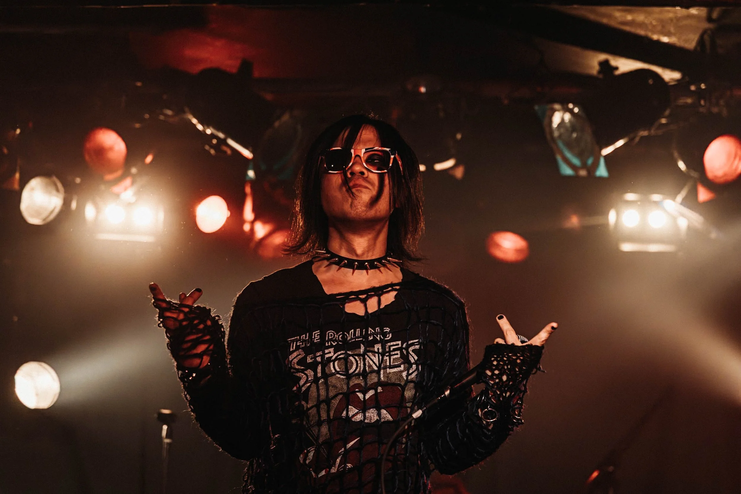 A rock musician on stage wearing sunglasses, a black ripped shirt with The Rolling Stones logo, a spiked choker, black fishnet gloves, and curly black hair, with stage lights and equipment in the background.