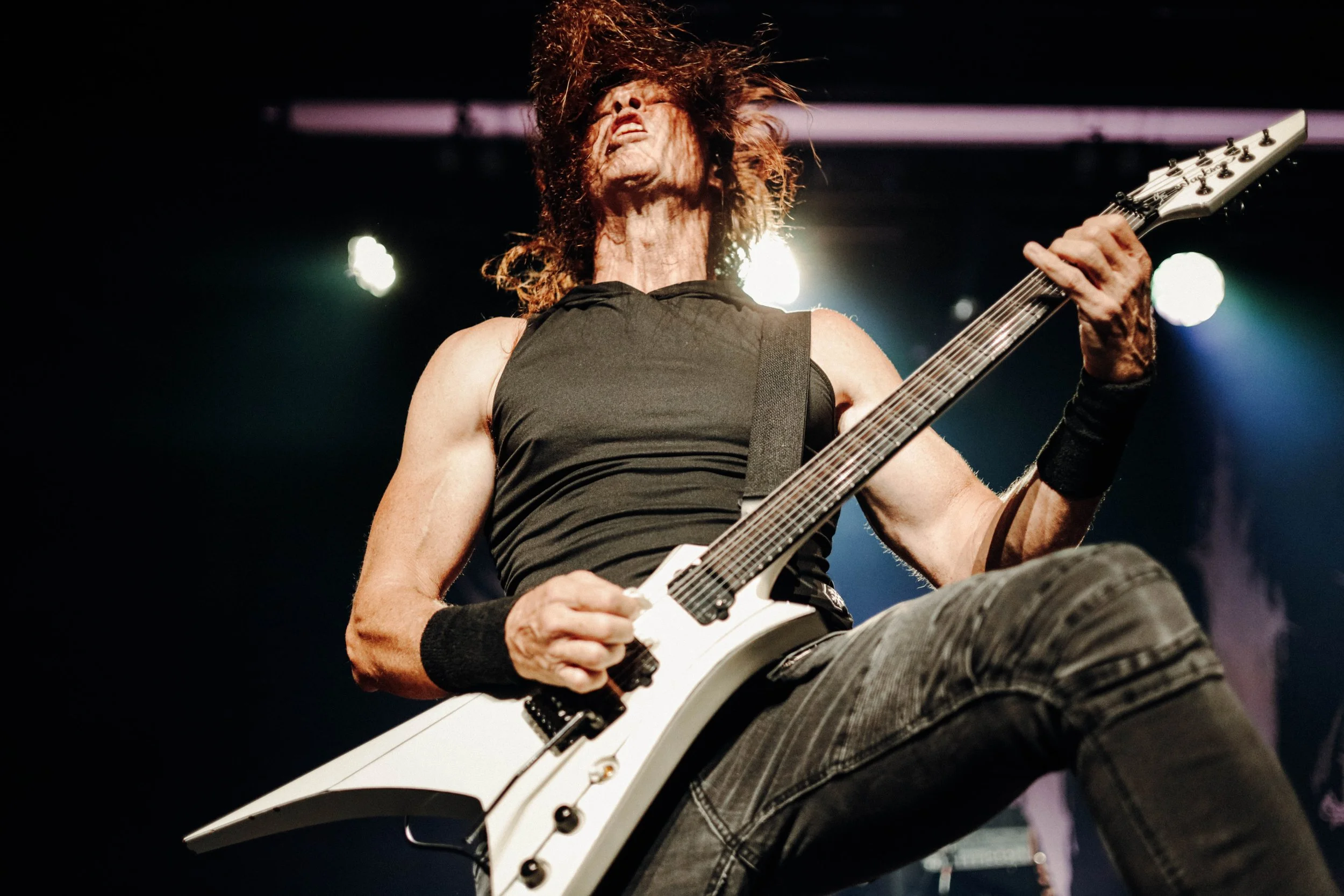 A guitarist performs on stage with long hair, wearing a black sleeveless shirt and black wristbands, playing an electric guitar with a pointed white body and black hardware, under stage lights.