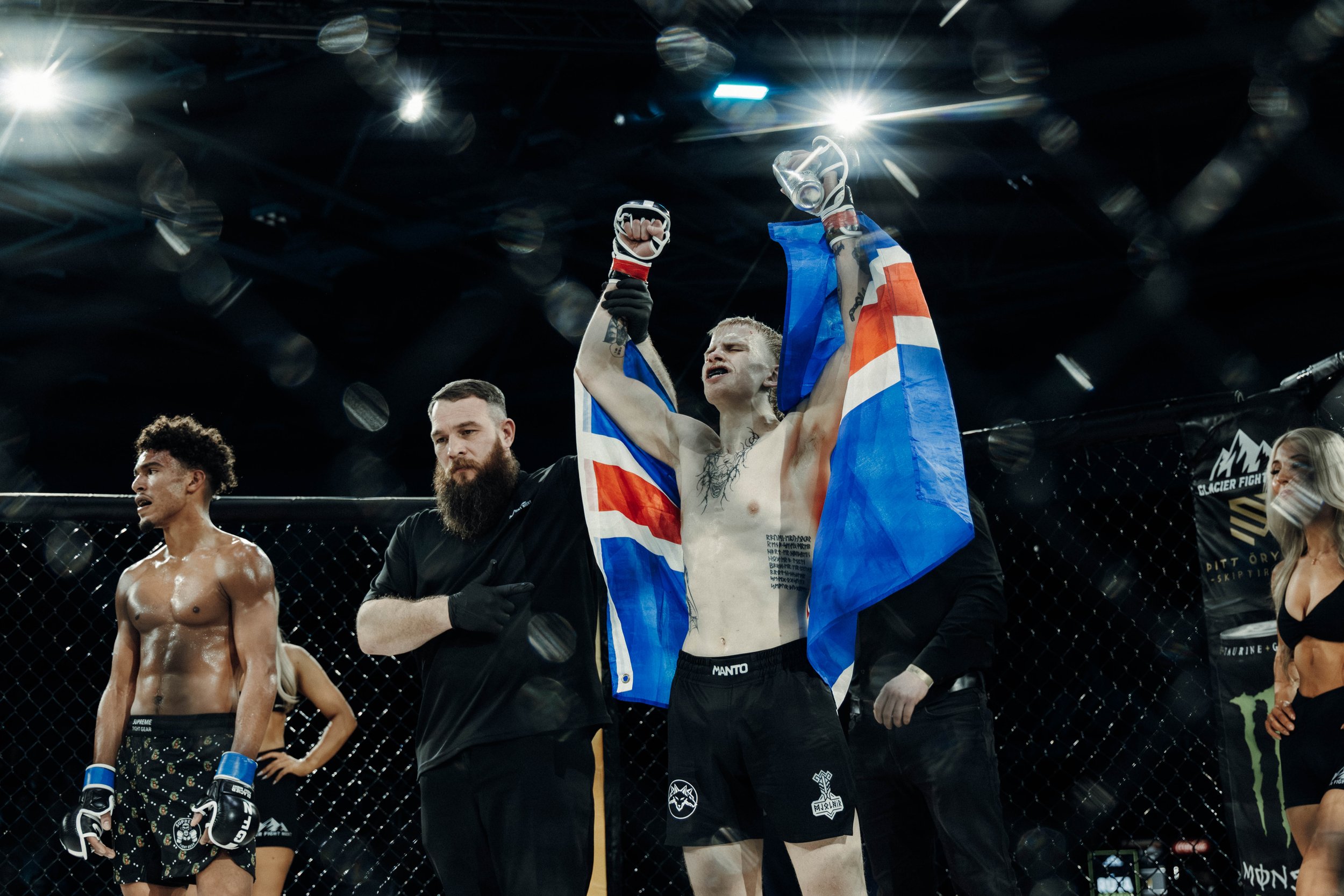 Mixed martial arts fighter with short blonde hair celebrating victory with arms raised, holding a flag over his shoulders inside octagon cage, with other fighters and officials around, under bright overhead lights.