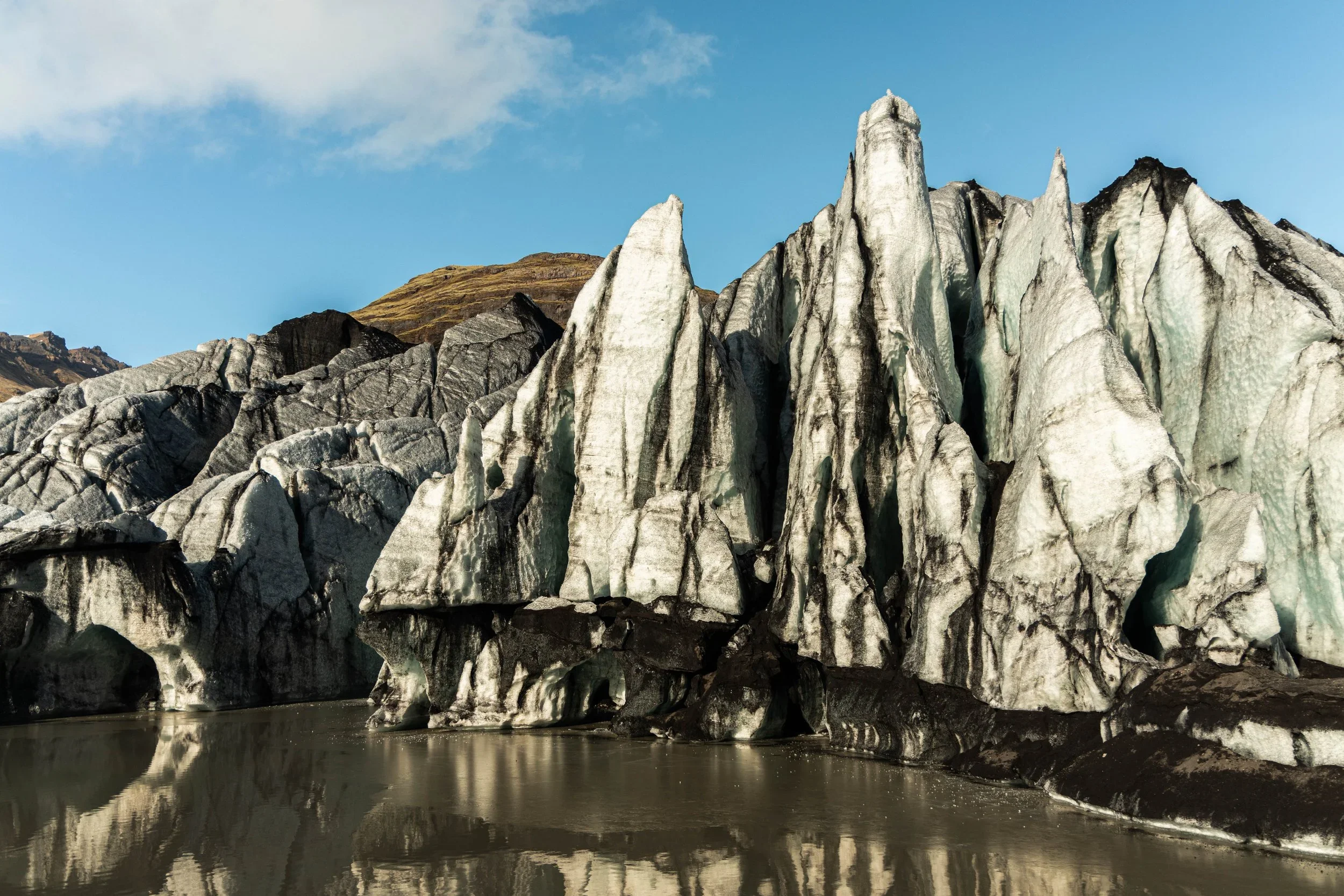 Large glacier with jagged ice formations reflecting in a still body of water, with a blue sky and distant mountain landscape in the background.