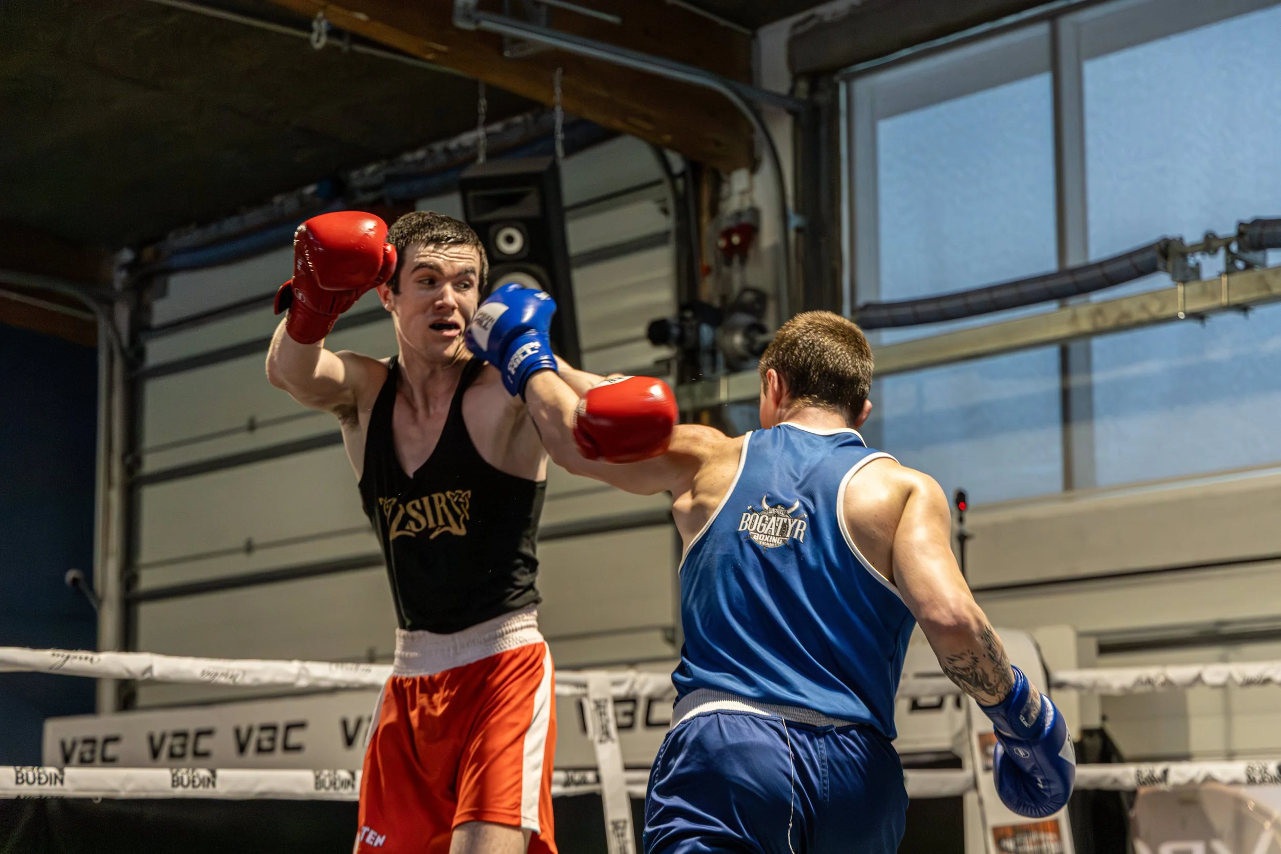 Two male boxers engaging in a match inside a boxing ring, one with red gloves and the other with blue gloves, with the boxer in black shorts throwing a punch.