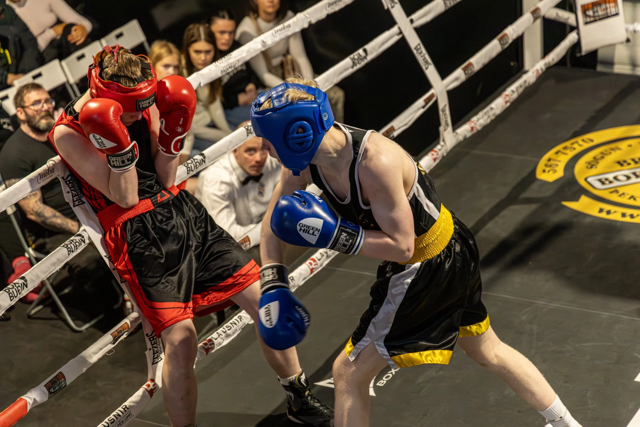 Two female boxers in boxing gear sparring inside a boxing ring, with one in red and the other in black and yellow, and spectators watching in the background.