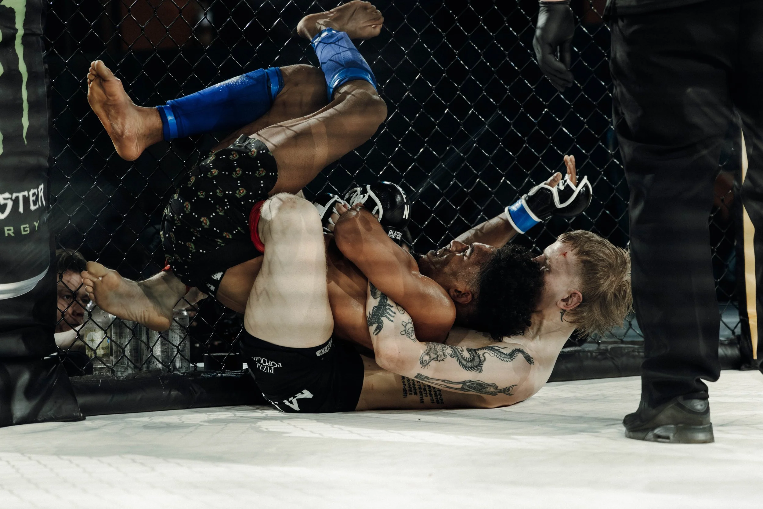 Two mixed martial artists grappling on the ground inside an octagon cage, with a referee nearby. The fighter on top is wearing black shorts with red and white accents and blue gloves, while the other fighter is shirtless with tattoos, wearing black s