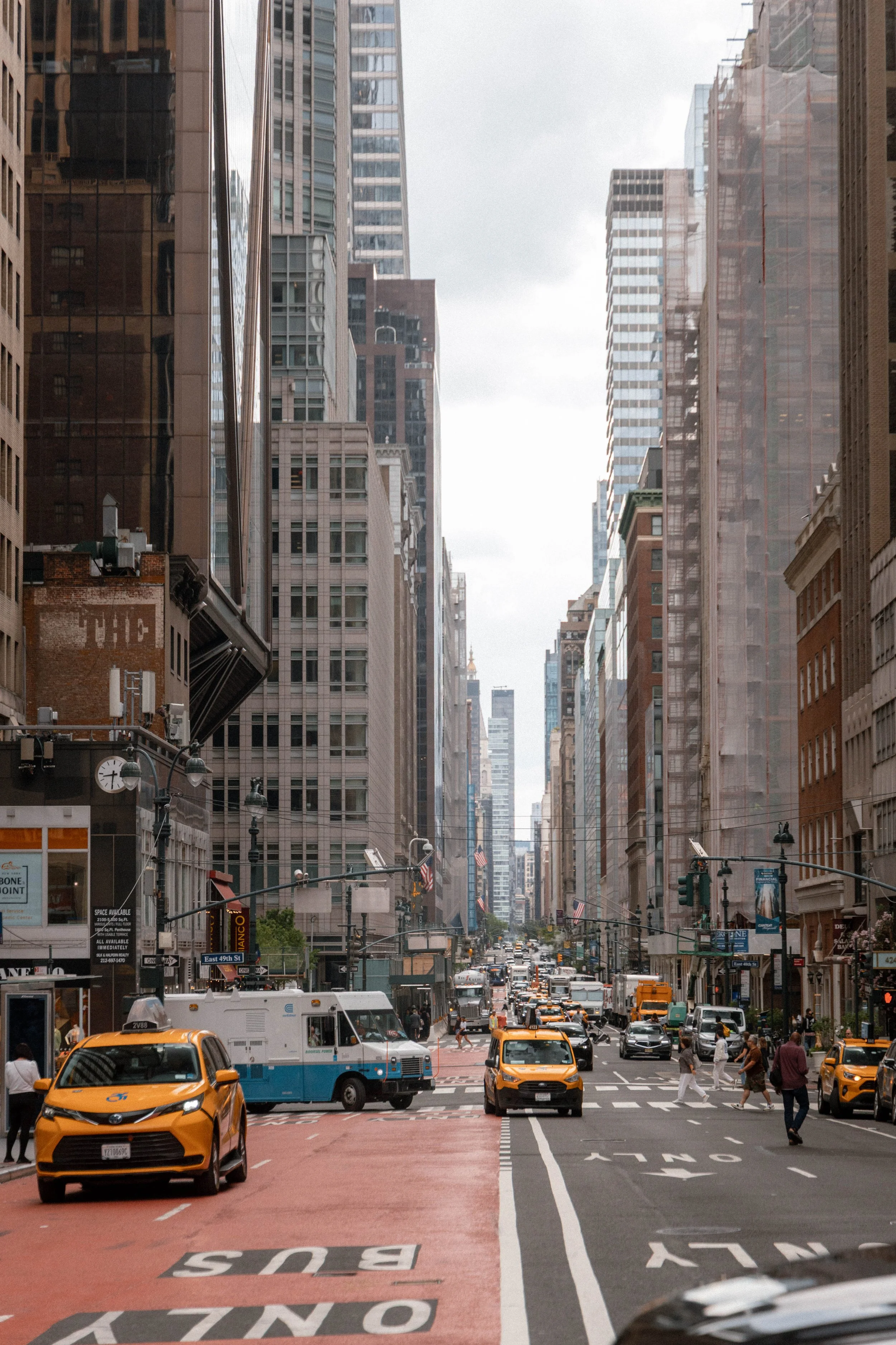 A busy city street with yellow taxis, pedestrians, and tall buildings on both sides under a cloudy sky.
