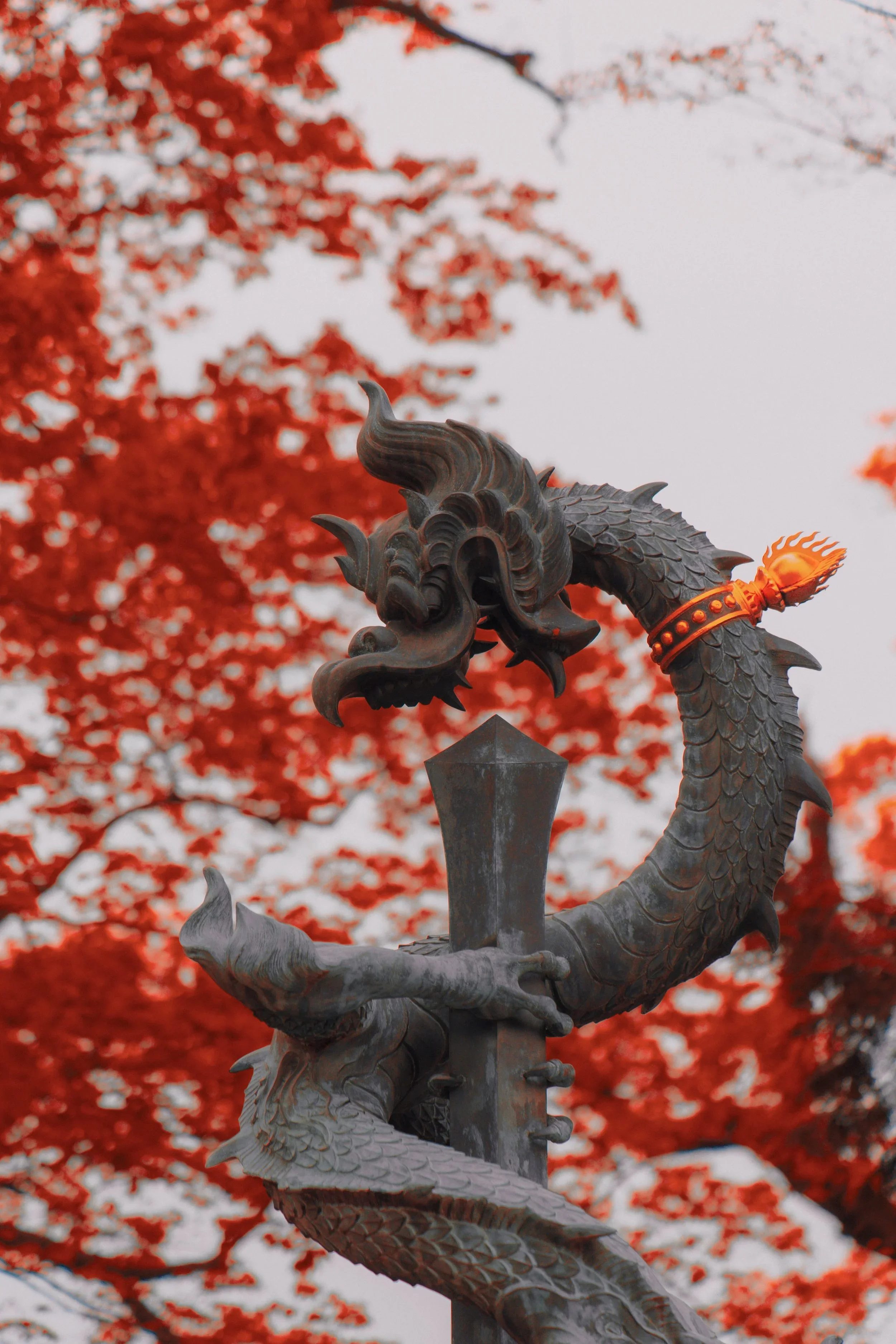 A decorative metal dragon sculpture with a fiery element around its neck, set against a backdrop of bright red autumn leaves.