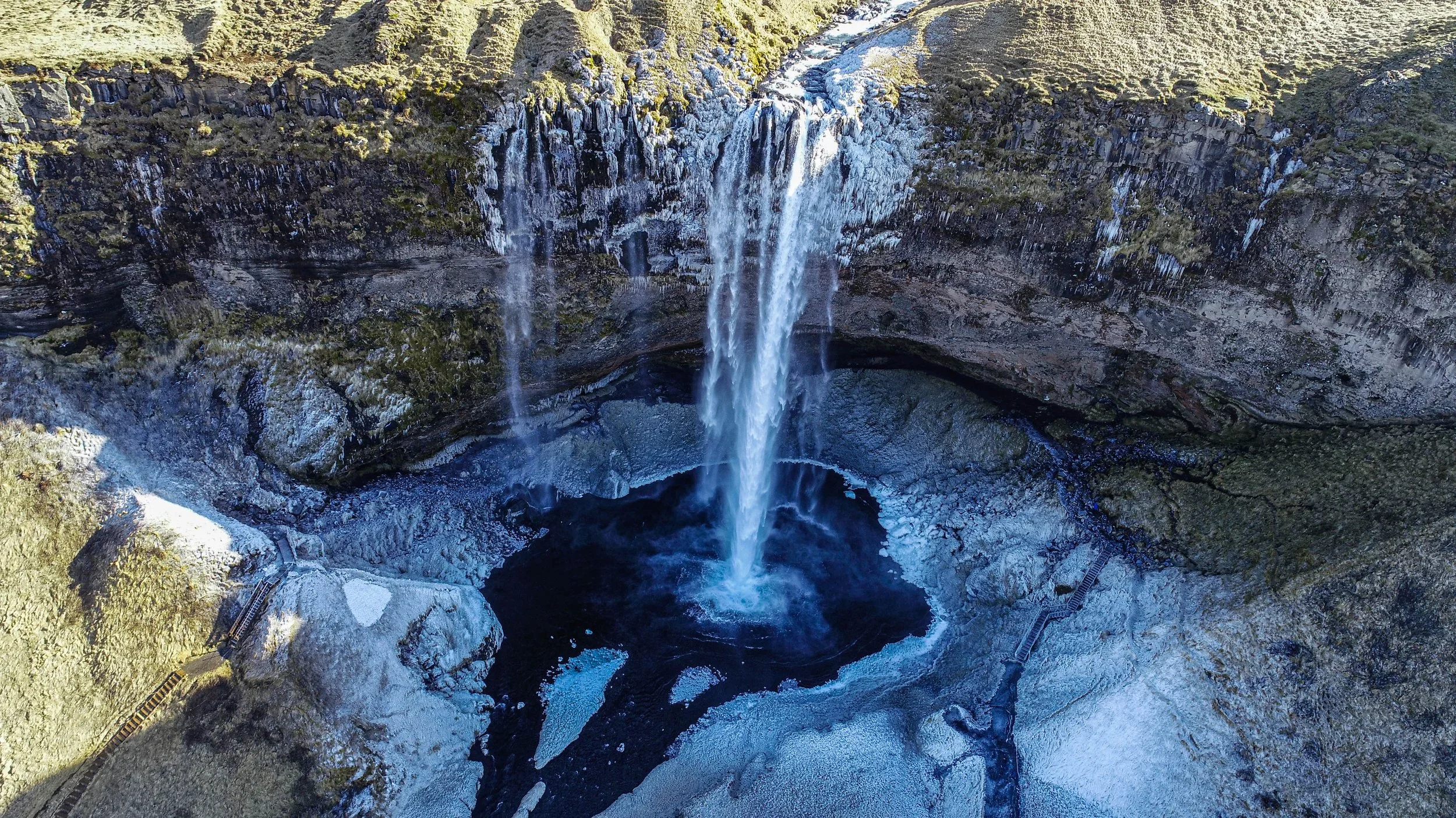 Aerial view of a tall waterfall cascading into a dark pool below, surrounded by rocky and mossy cliffs.