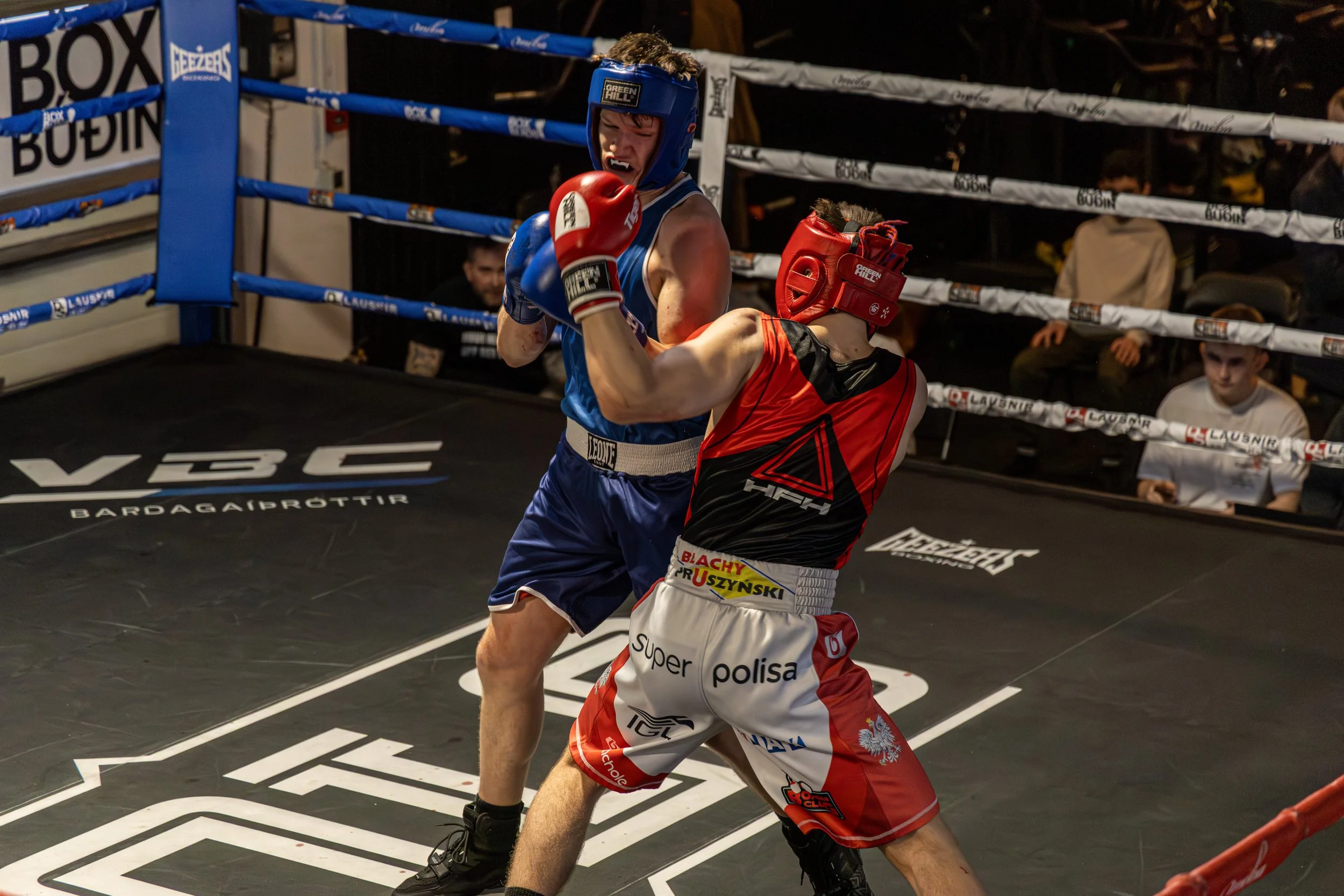 Two male boxers in a match, one wearing a blue uniform and headgear, the other in red, fighting inside a boxing ring with rope barriers and an audience watching.
