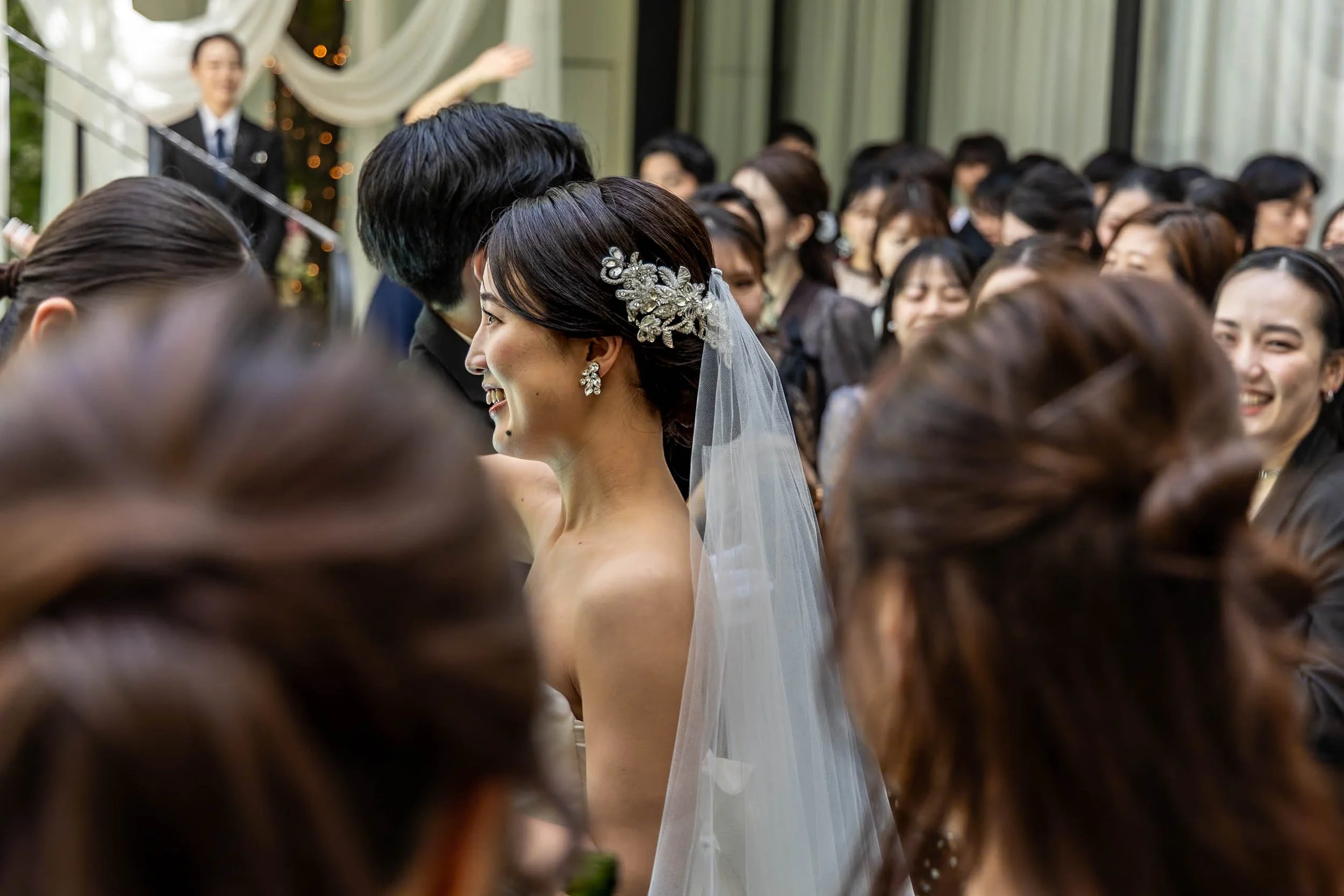 A bride smiling at a wedding reception, surrounded by guests in an indoor event space.