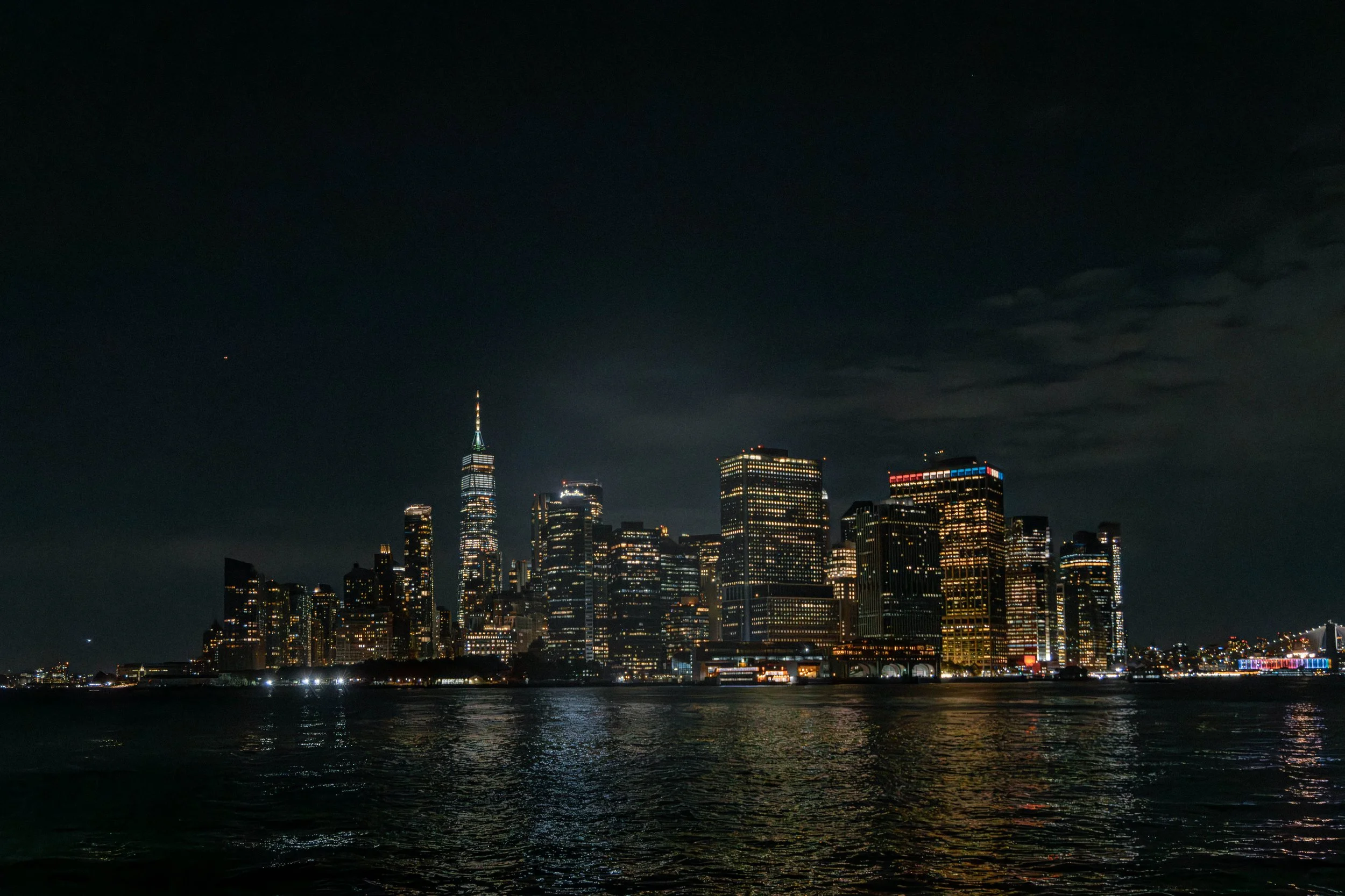 Nighttime city skyline of Manhattan with illuminated skyscrapers, including the Empire State Building, reflected on dark water.