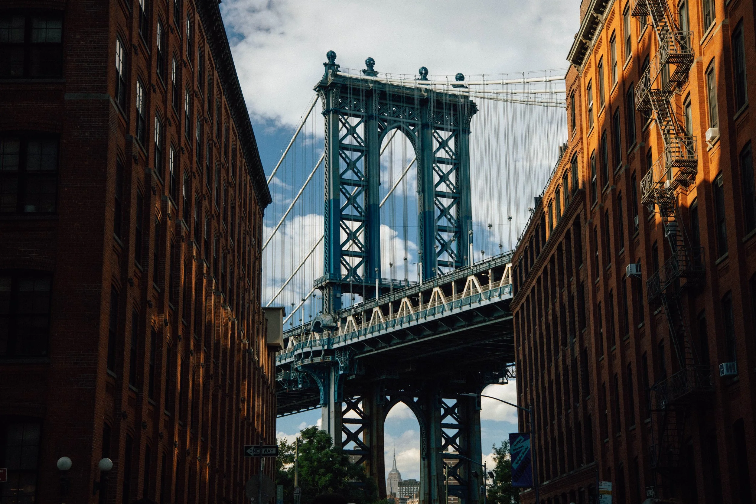 View of the Manhattan Bridge in New York City, seen through old brick buildings on a partly cloudy day.
