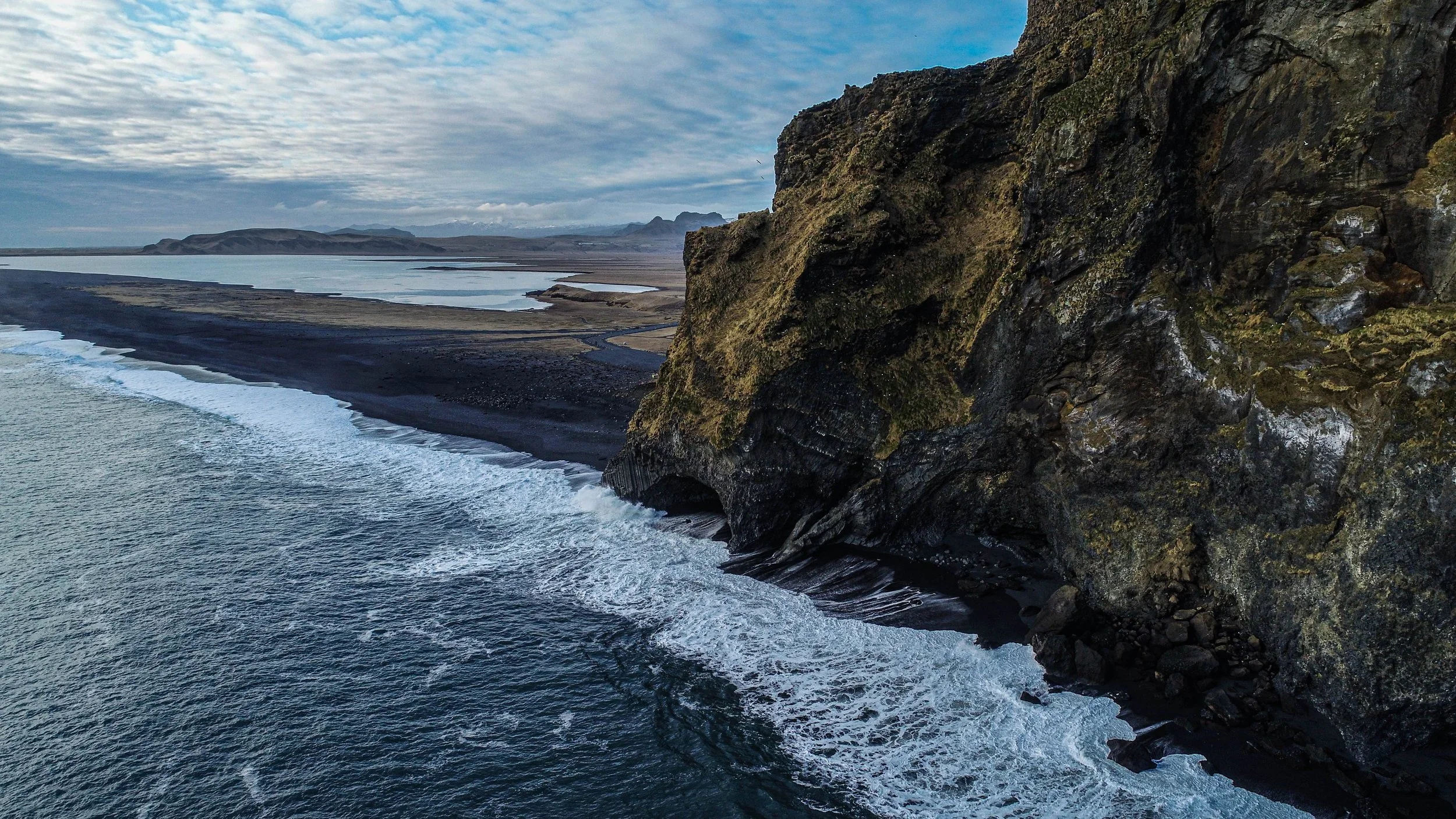 A rugged coastline with a large dark cliff on the right, crashing waves at the base, and a flat plain with a lake and mountains in the background under a cloudy sky.