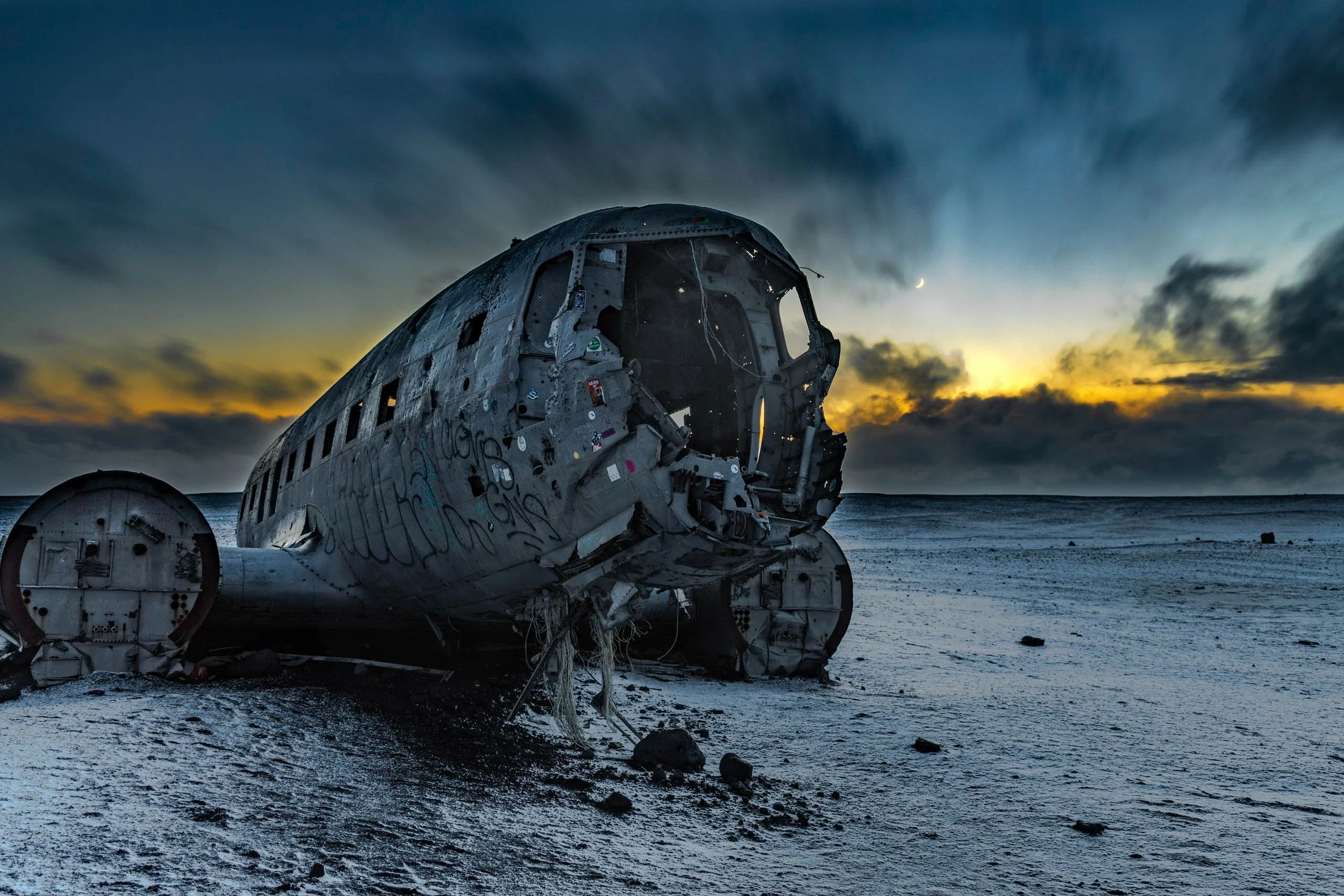 Remains of a destroyed airplane lying on a desert landscape at sunset, with a dramatic sky and clouds in the background.
