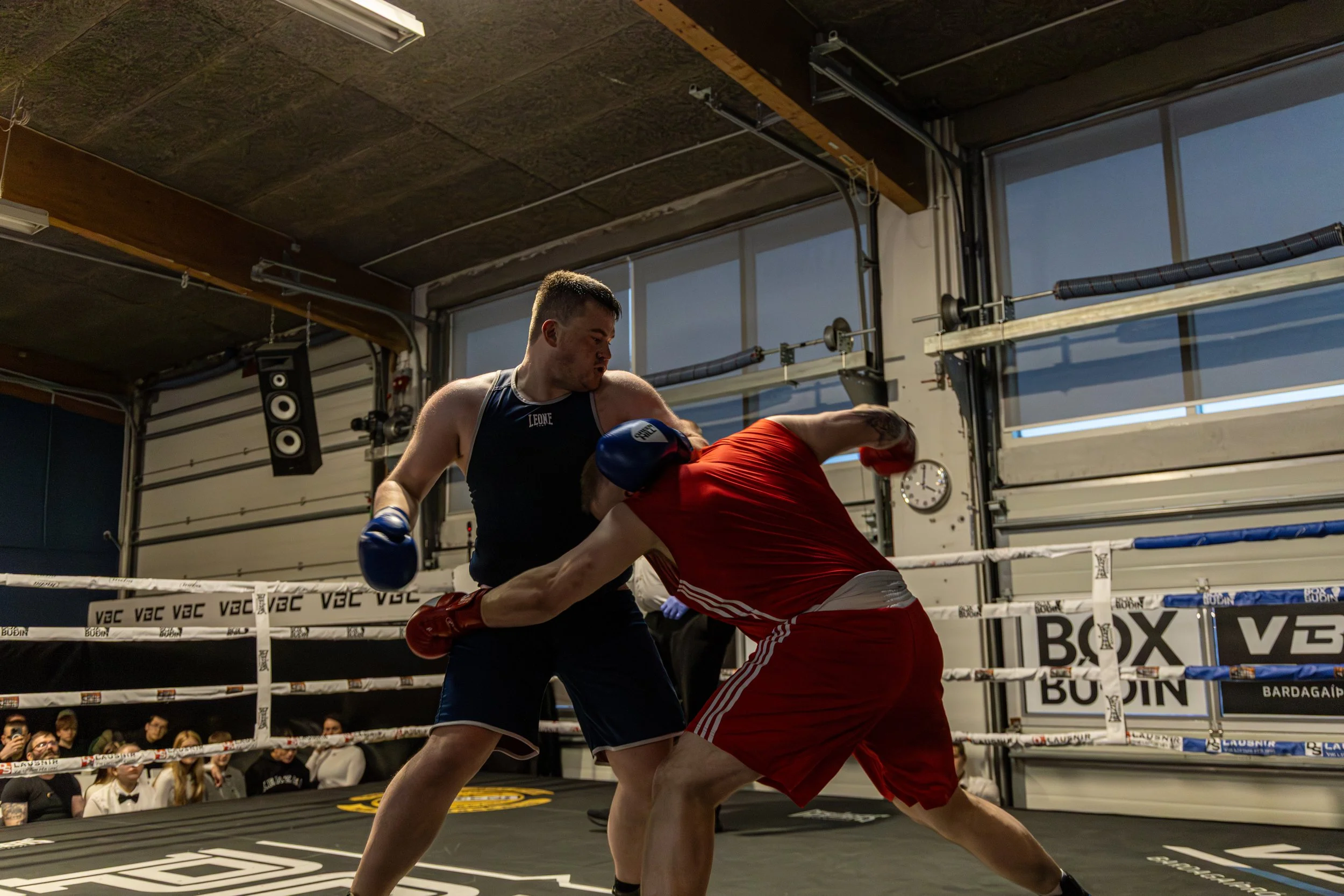 Two men engaged in a boxing match inside a gym ring, with an audience watching.