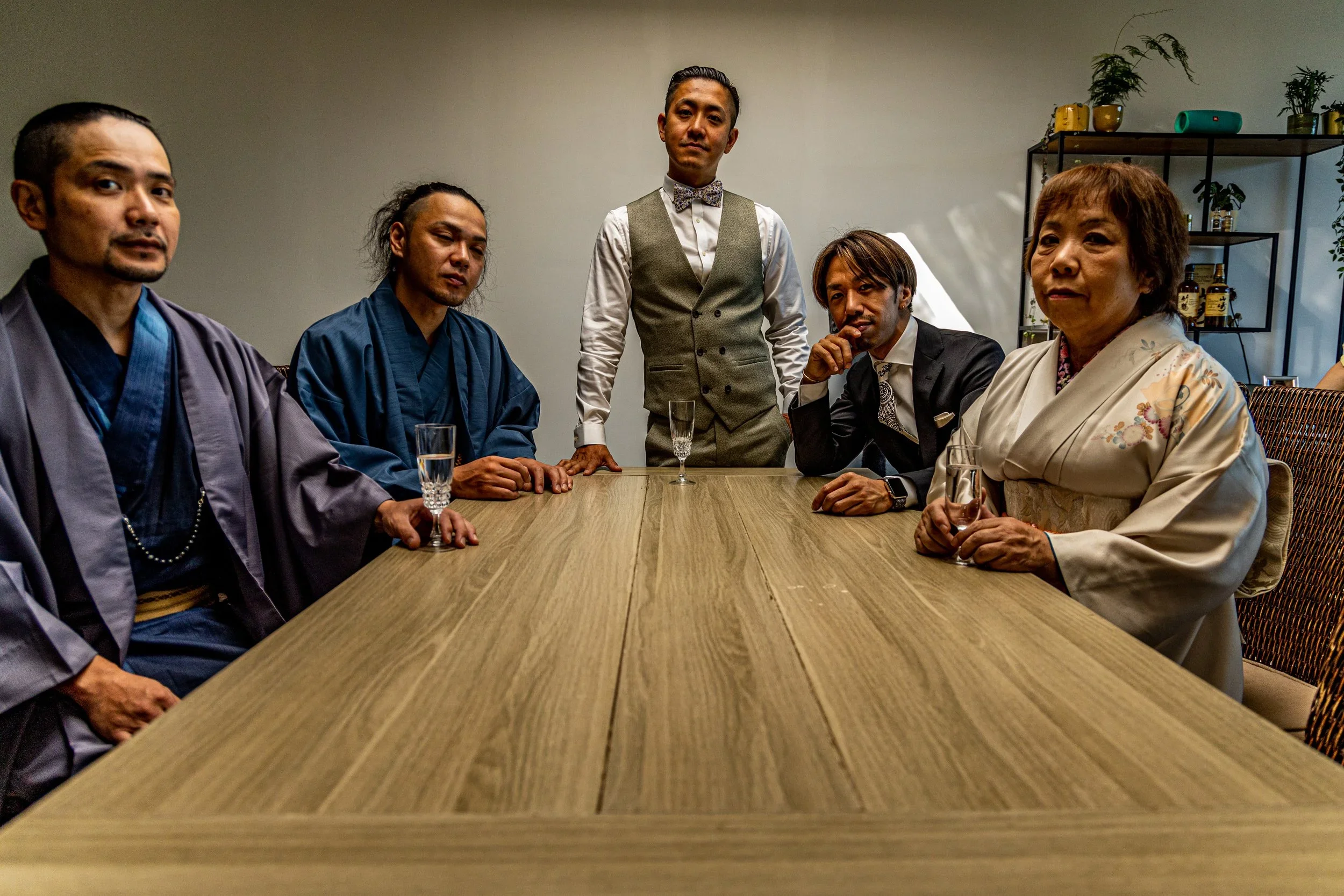 A group of six people around a wooden table, with five seated and one standing behind. The seated individuals include women in traditional kimonos and men in suits, holding glasses of champagne. The standing man is dressed in a vest, shirt, and bowti