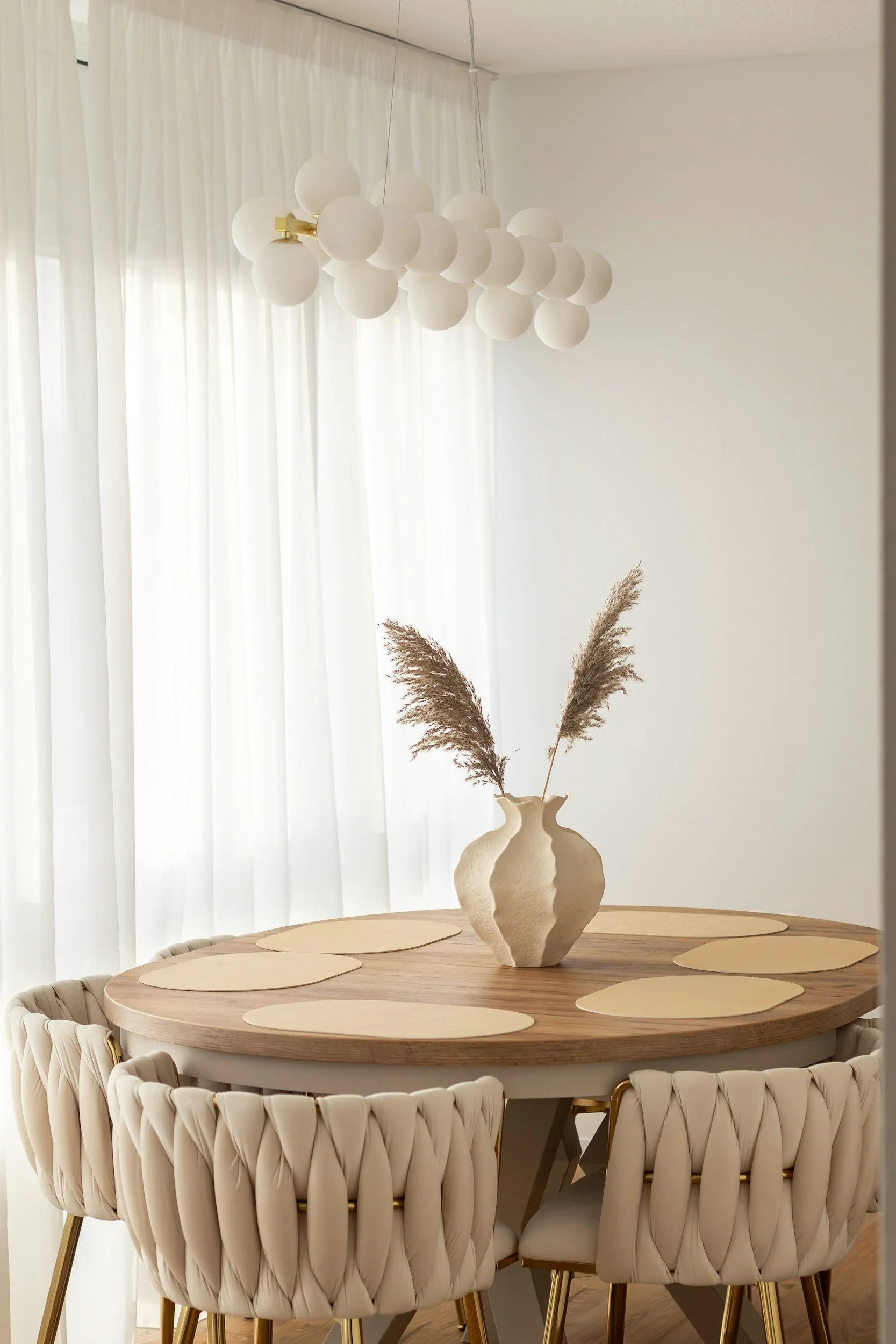 A round wooden dining table with six beige woven chairs, a tall beige vase with dried pampas grass, a white modern chandelier with multiple globes, and white curtains.