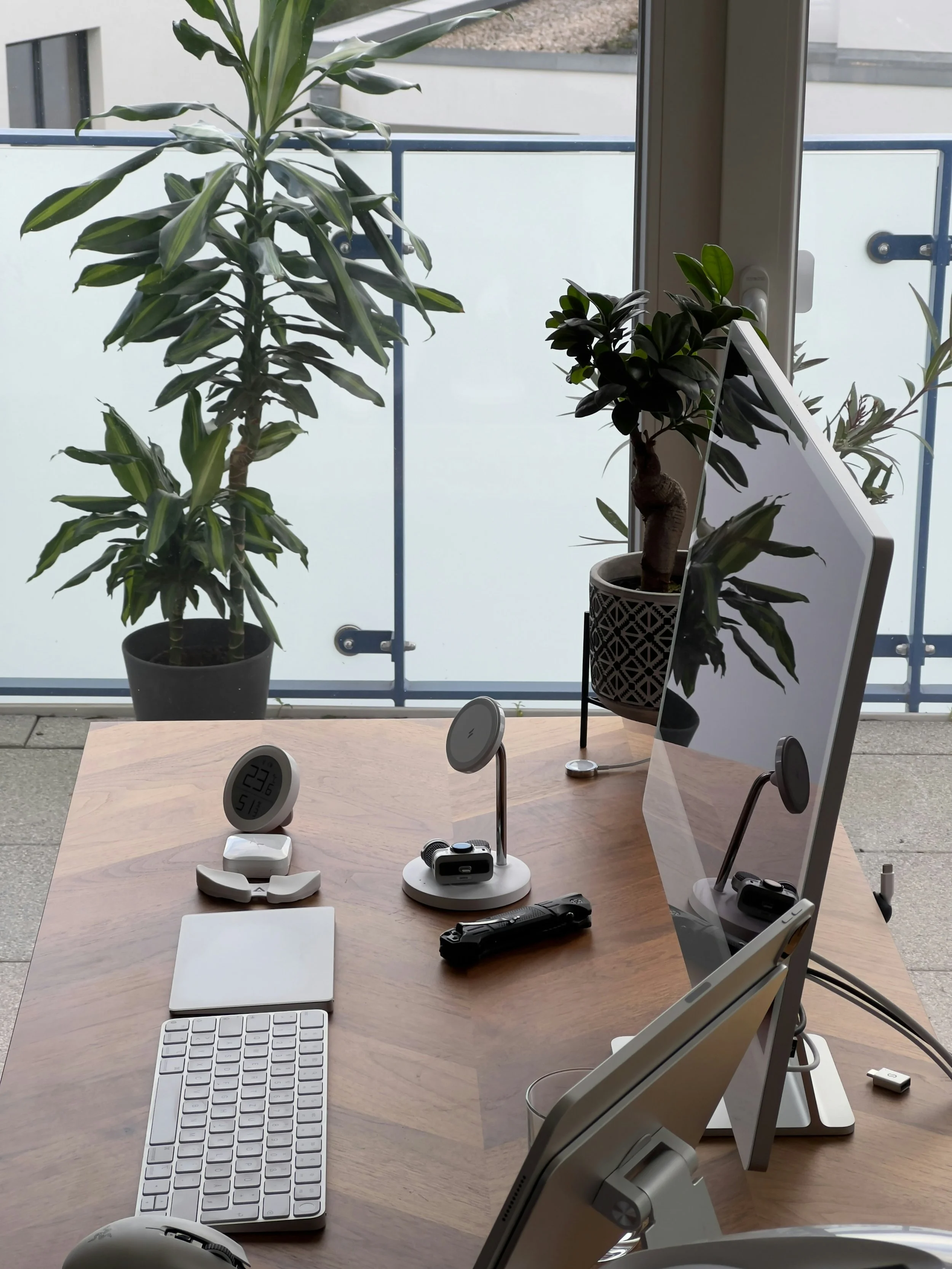 Modern office desk with a computer monitor, wireless keyboard, white mouse, and various electronic gadgets. Two potted plants are on the desk, and in the background, there are large green plants outside a glass window and balcony railing.