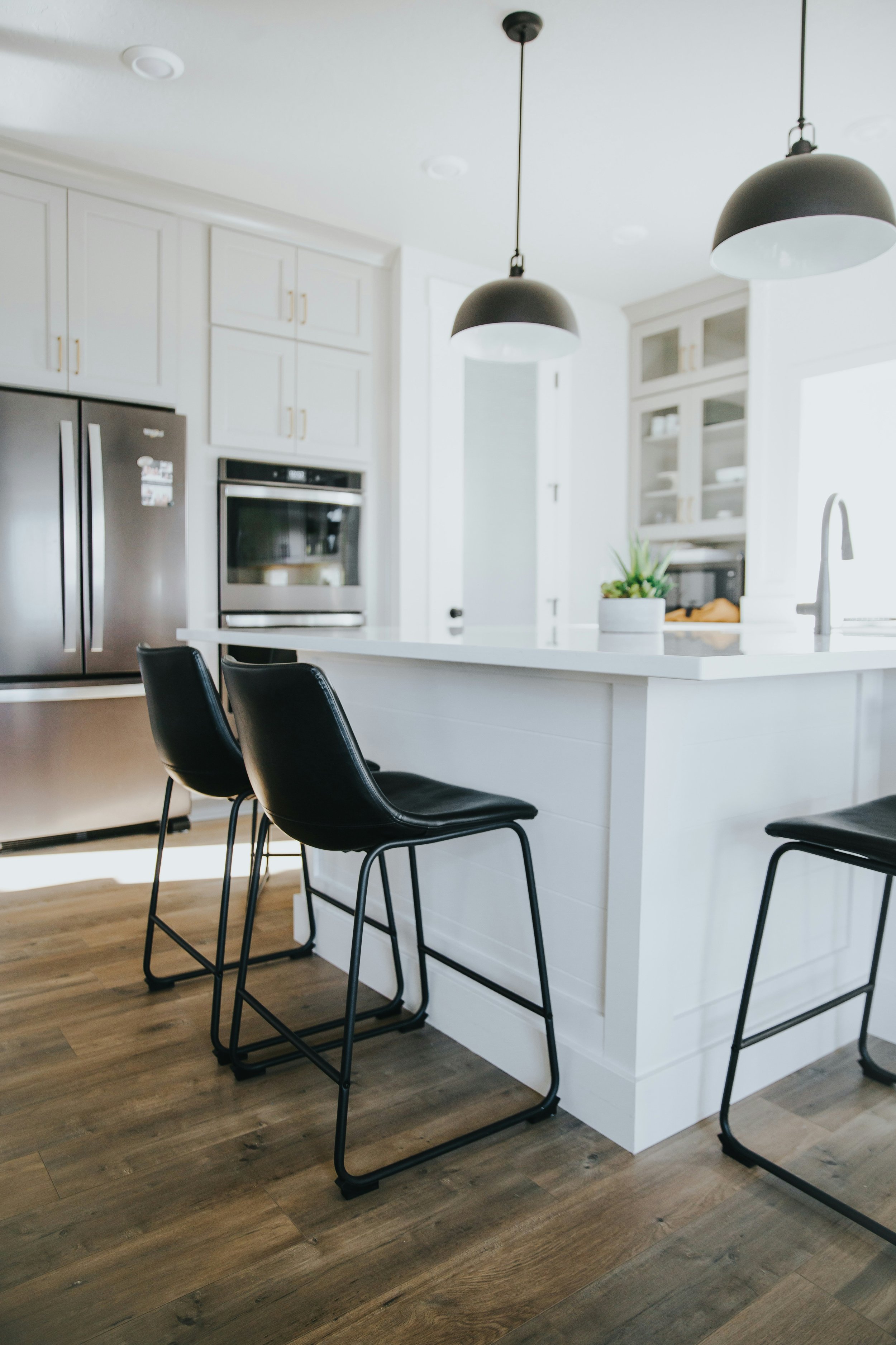 Modern kitchen with white cabinetry, black barstools, pendant lights, stainless steel refrigerator, microwave, and a countertop with a potted plant.