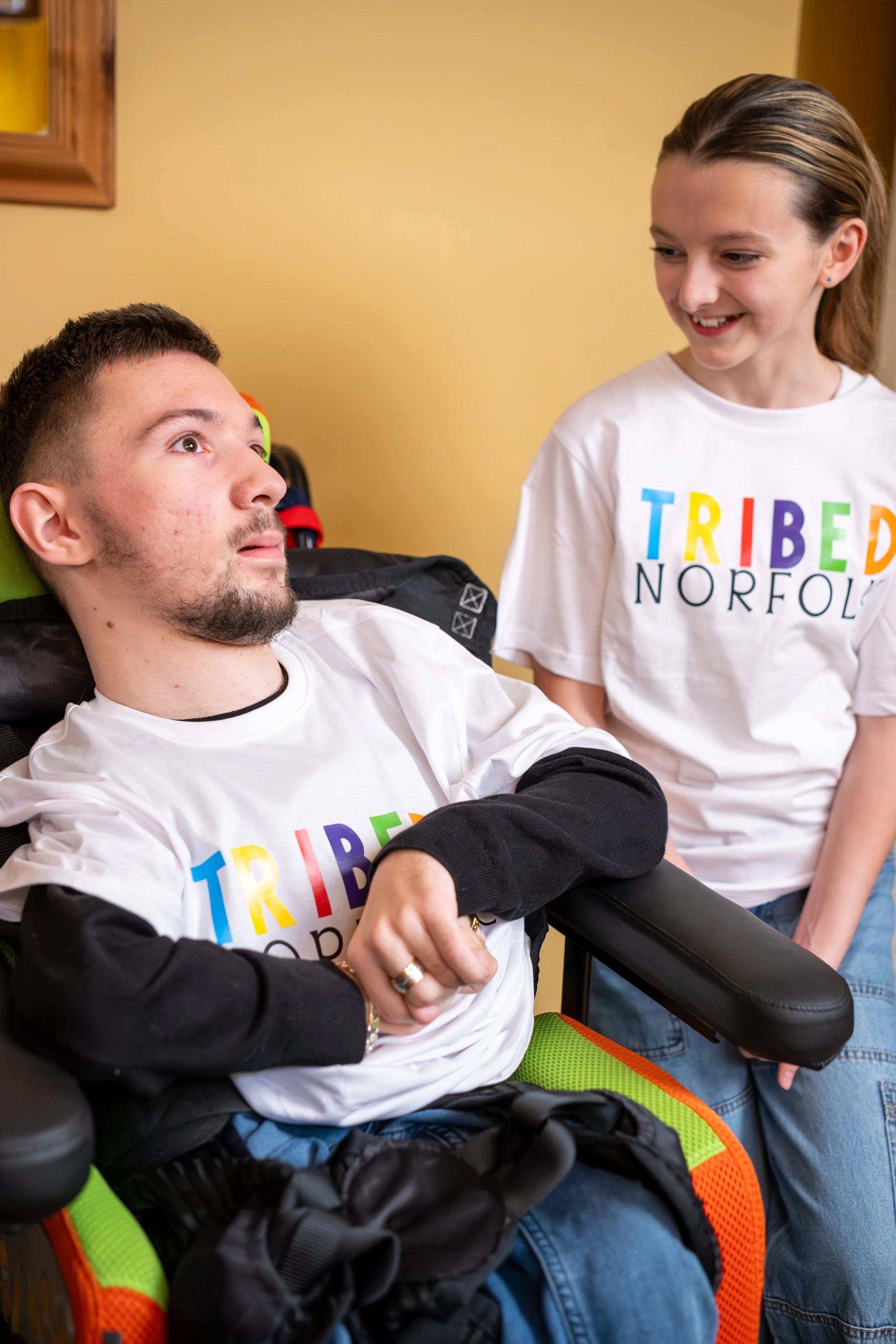 A young man in a wheelchair with a black jacket, and a young girl smiling, both wearing white T-shirts with the colorful word 'TRIBED' on them, indoor setting with a yellow wall.
