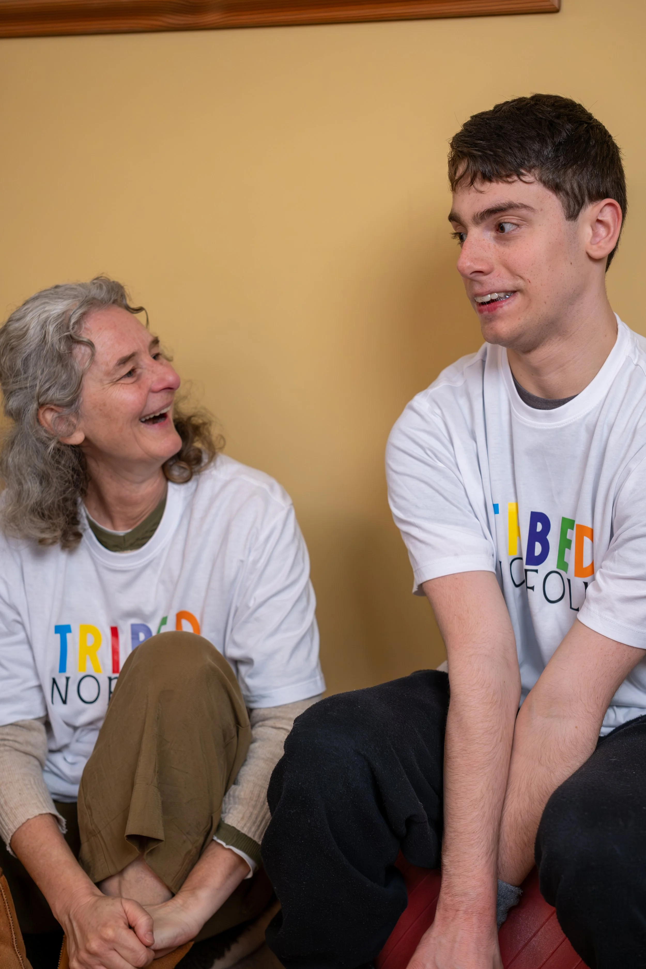 A young man with short dark hair and light skin is talking to an older woman with curly gray hair and light skin, both wearing white T-shirts with rainbow-colored text. They are sitting close together indoors against a beige wall, smiling and enjoying their conversation.