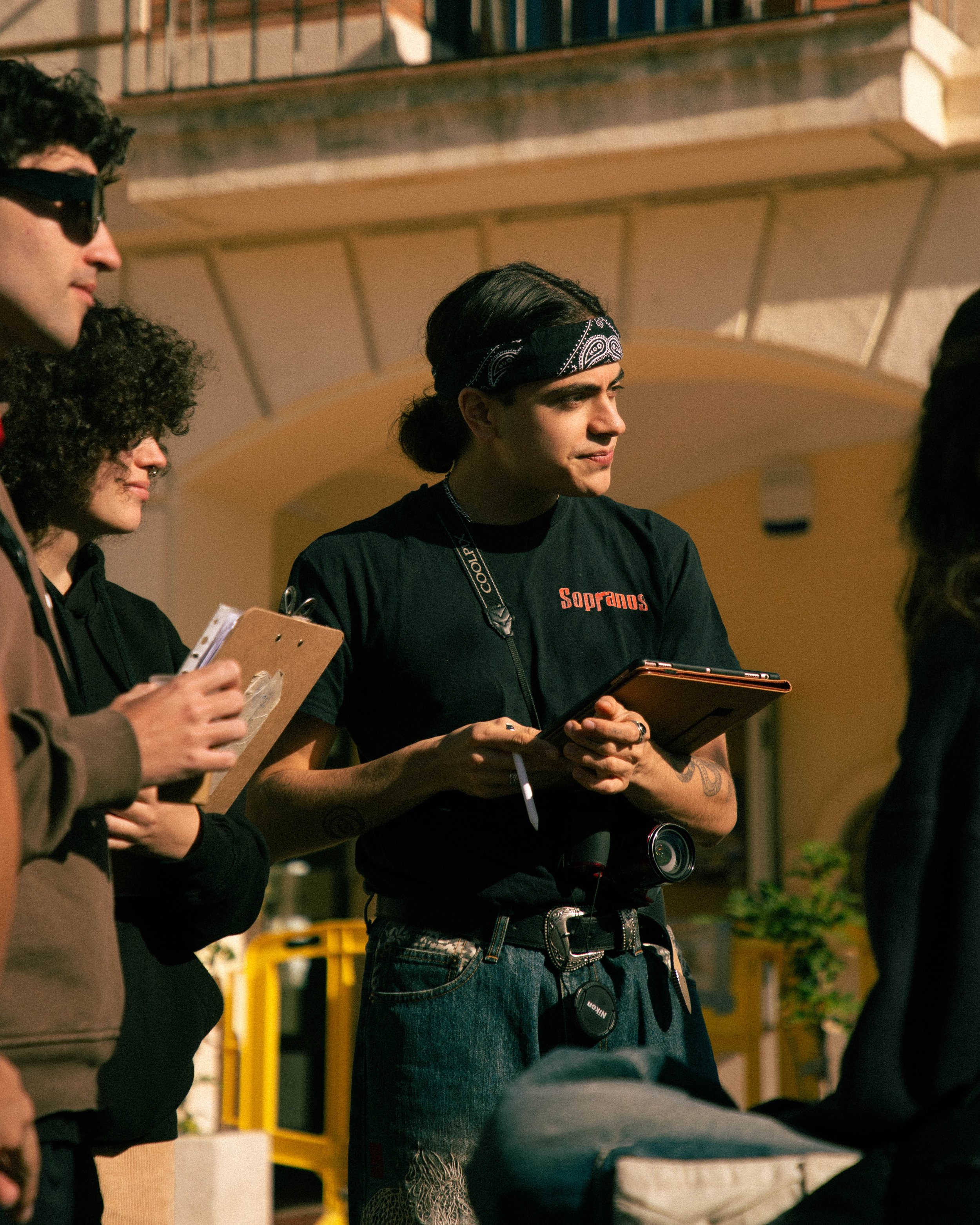 Grupo de personas en una reunión al aire libre, una de ellas lleva una camiseta negra con la palabra 'Sopranos' y una bandana en la cabeza, sosteniendo una tableta y una libreta.