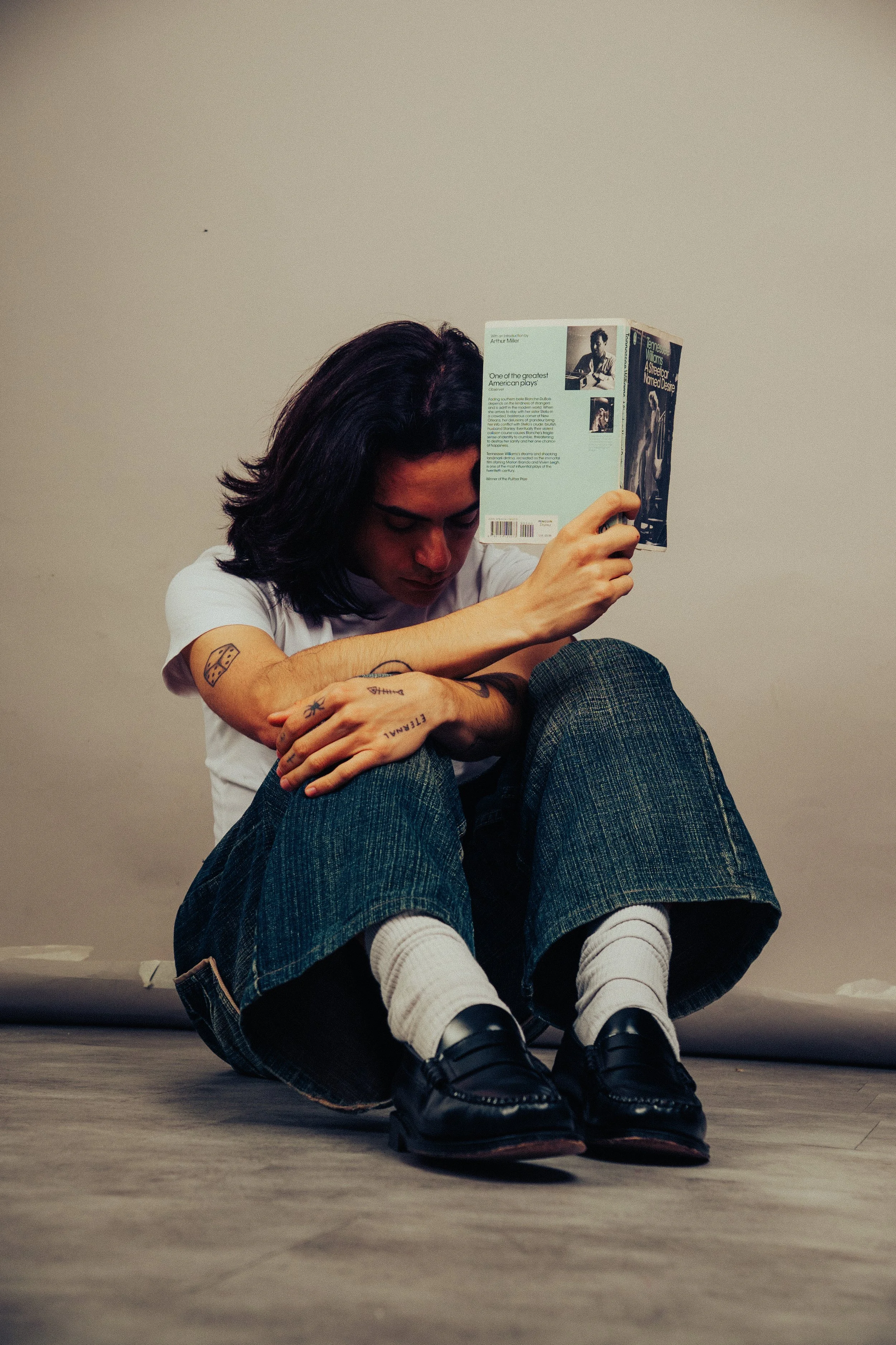 Persona sentada en el suelo, leyendo un libro, con cabello largo y oscuro, usando camiseta blanca, pantalones de mezclilla y zapatos negros con medias blancas.