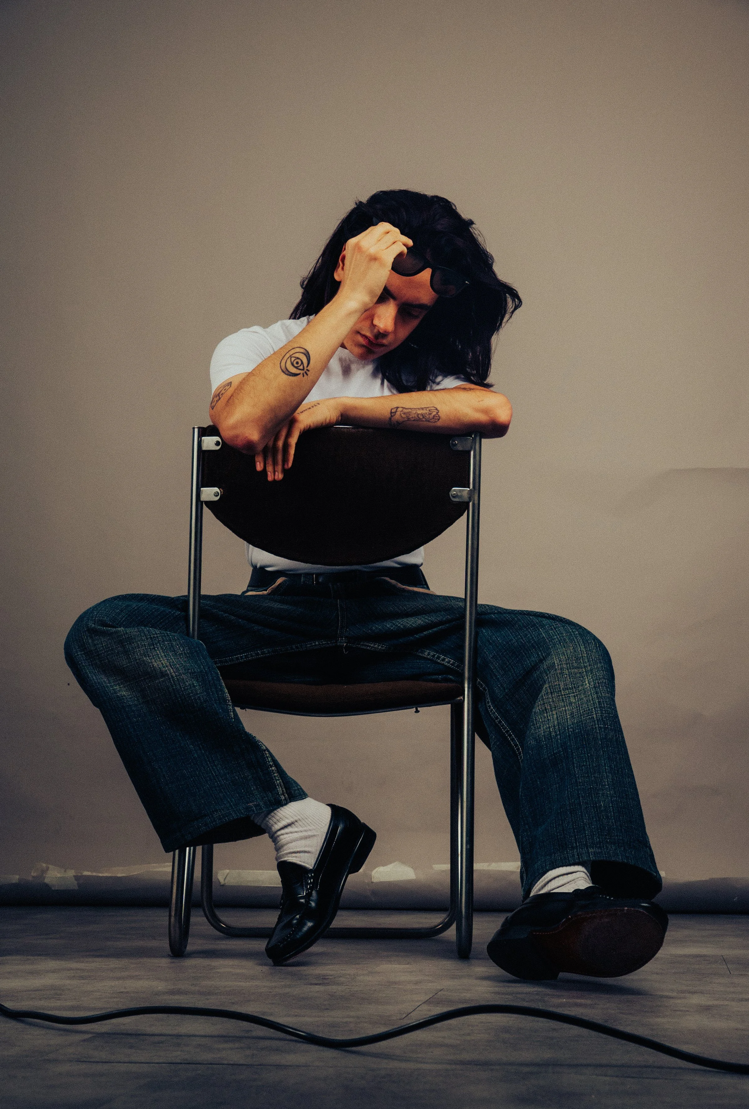 Joven sentado en una silla, con expresión pensativa, tocándose la cabeza y con tatuajes en sus brazos, vestimenta casual y fondo simple.