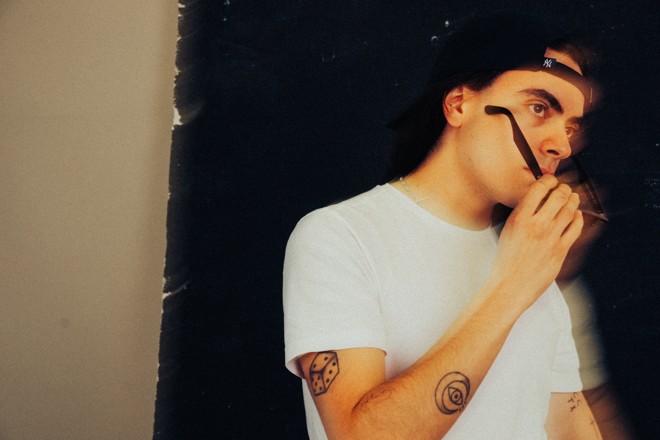 Joven con camiseta blanca, tatuajes en brazos, sosteniendo gafas delante de la cara, con fondo negro y beige.