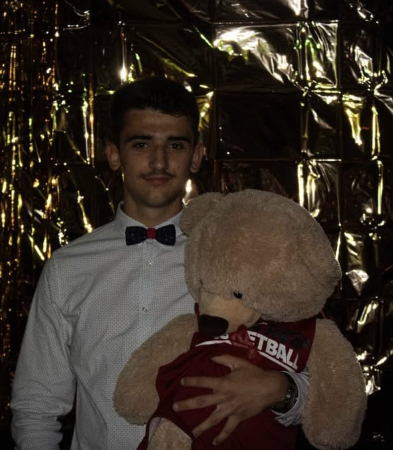 Jeune homme habillé en chemise blanche avec noeud papillon noir, tenant un grand ours en peluche beige portant un sweat rouge, devant un décor noir brillant et doré.